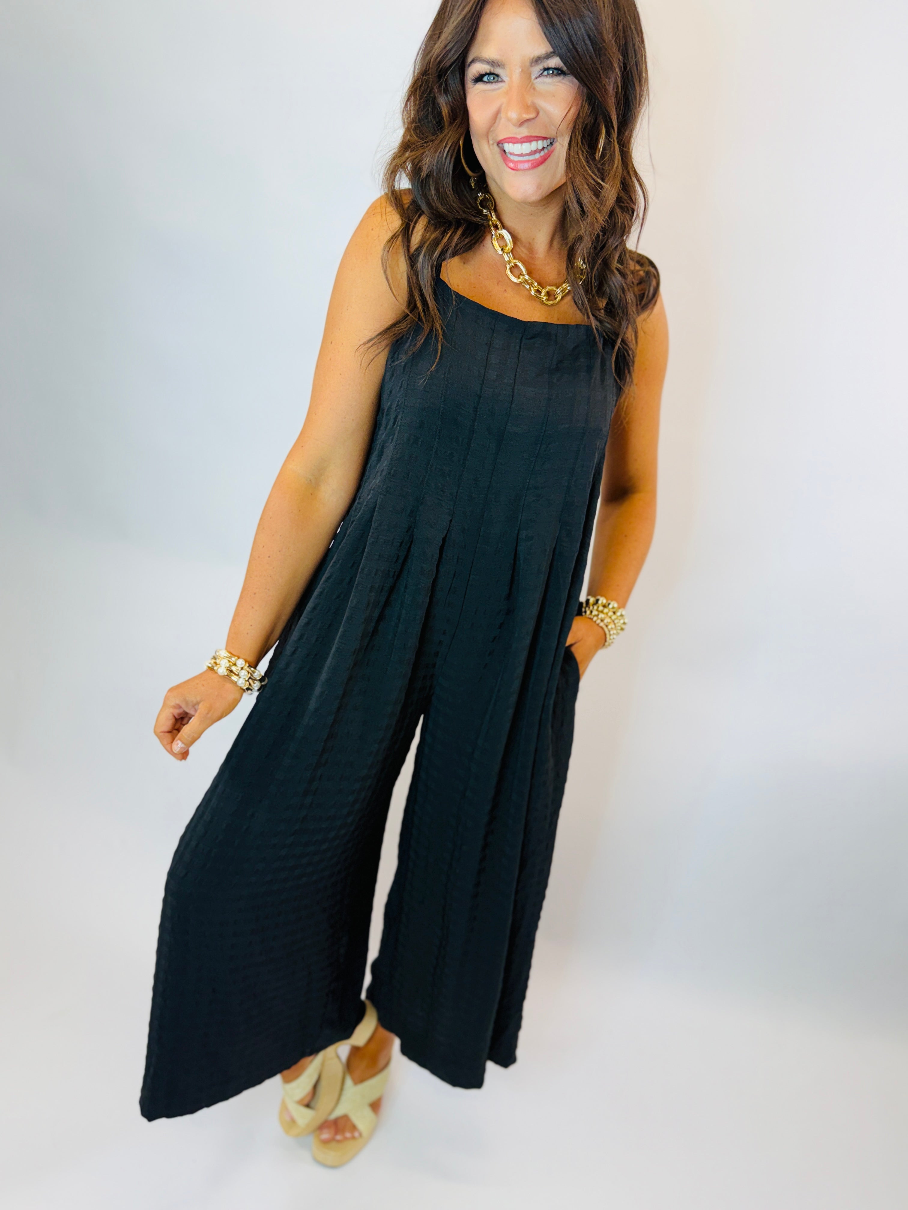 Woman wearing black sleeveless wide-leg jumpsuit with gold chain necklace and bracelets, styled with beige wedge sandals.