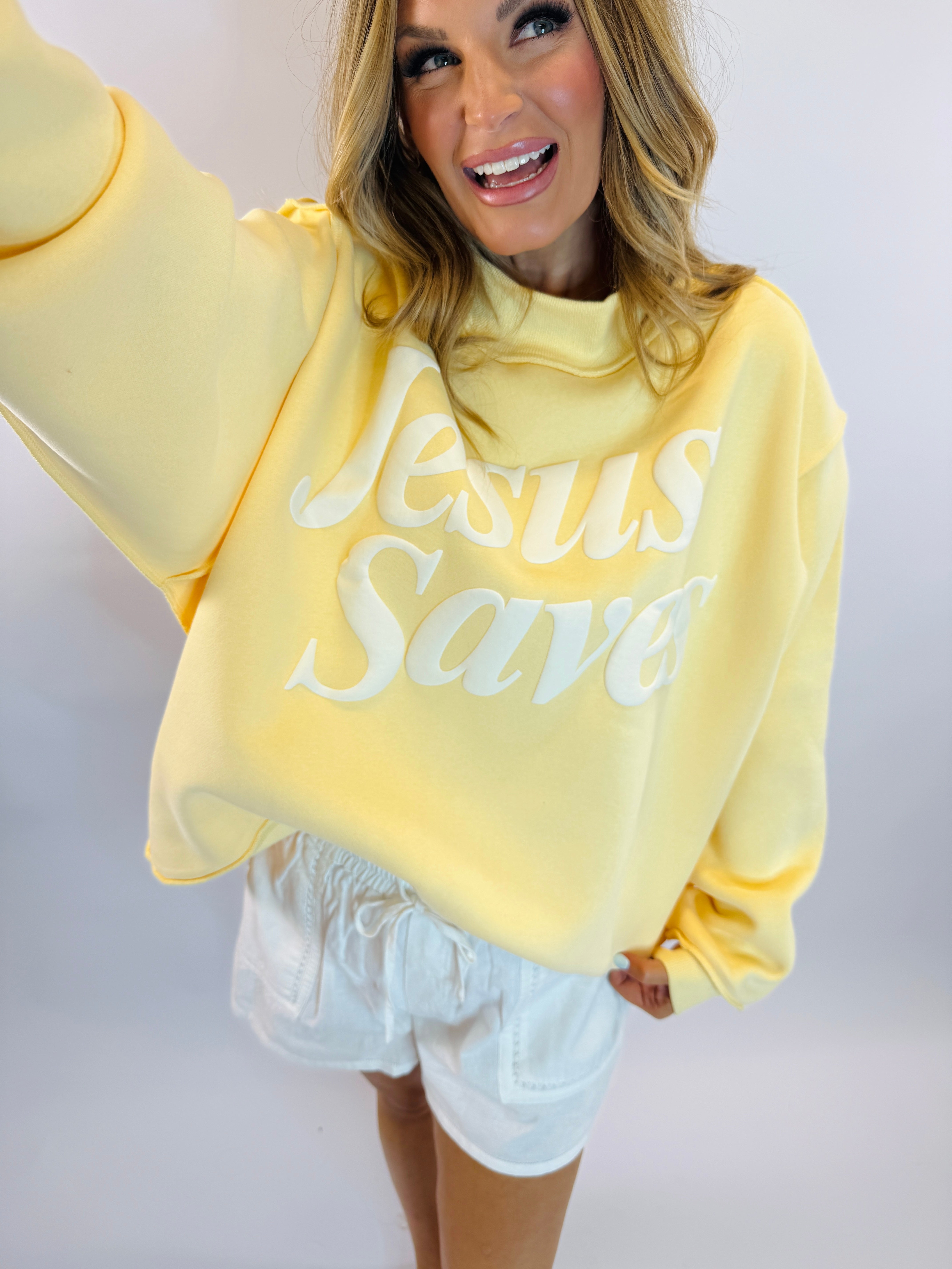 Woman wearing yellow mock neck sweatshirt with "Jesus Saves" text and white shorts, smiling with arms raised.