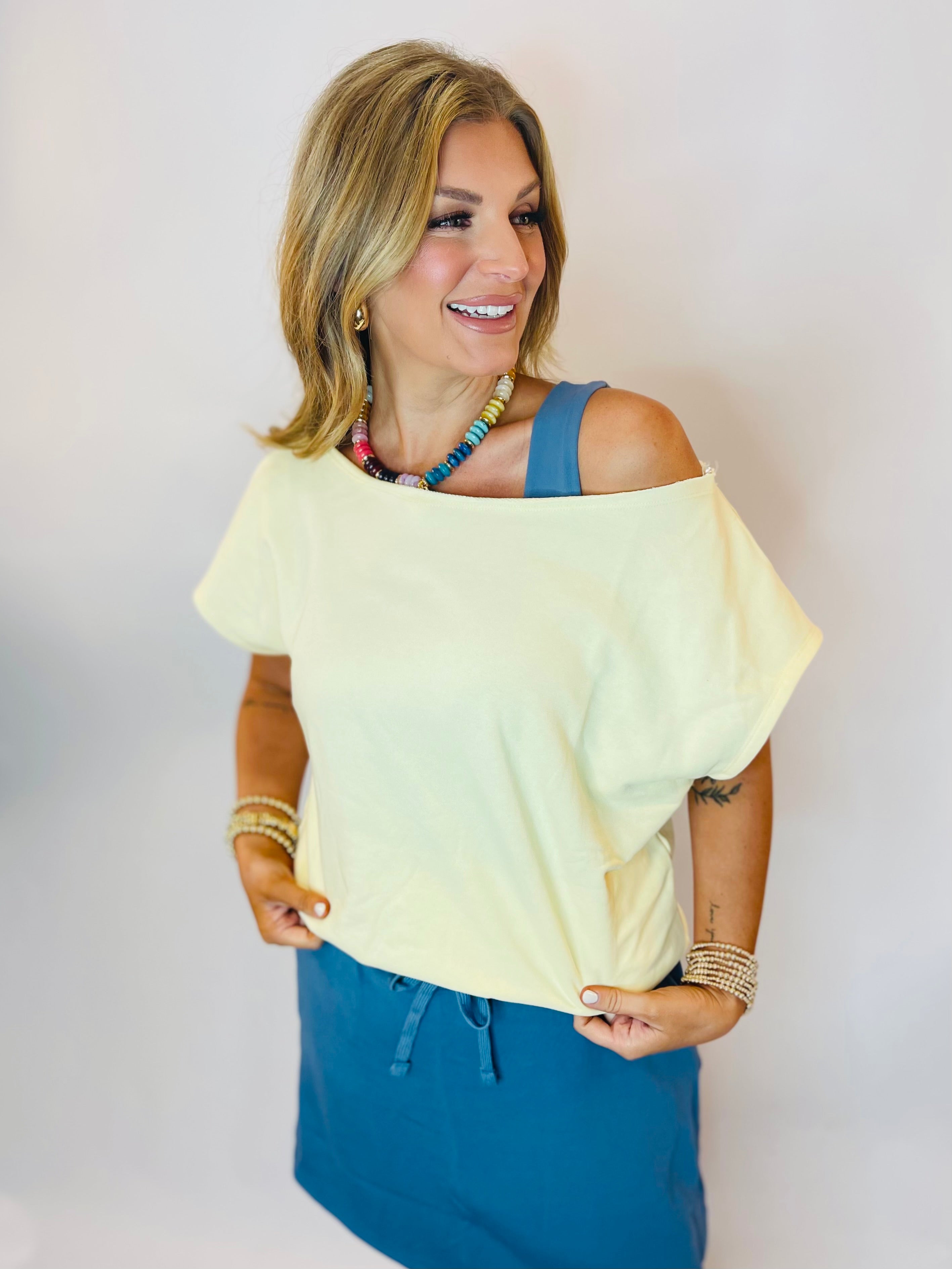 Woman wearing a loose lemon yellow off-shoulder top paired with a teal skirt and colorful beaded necklace.