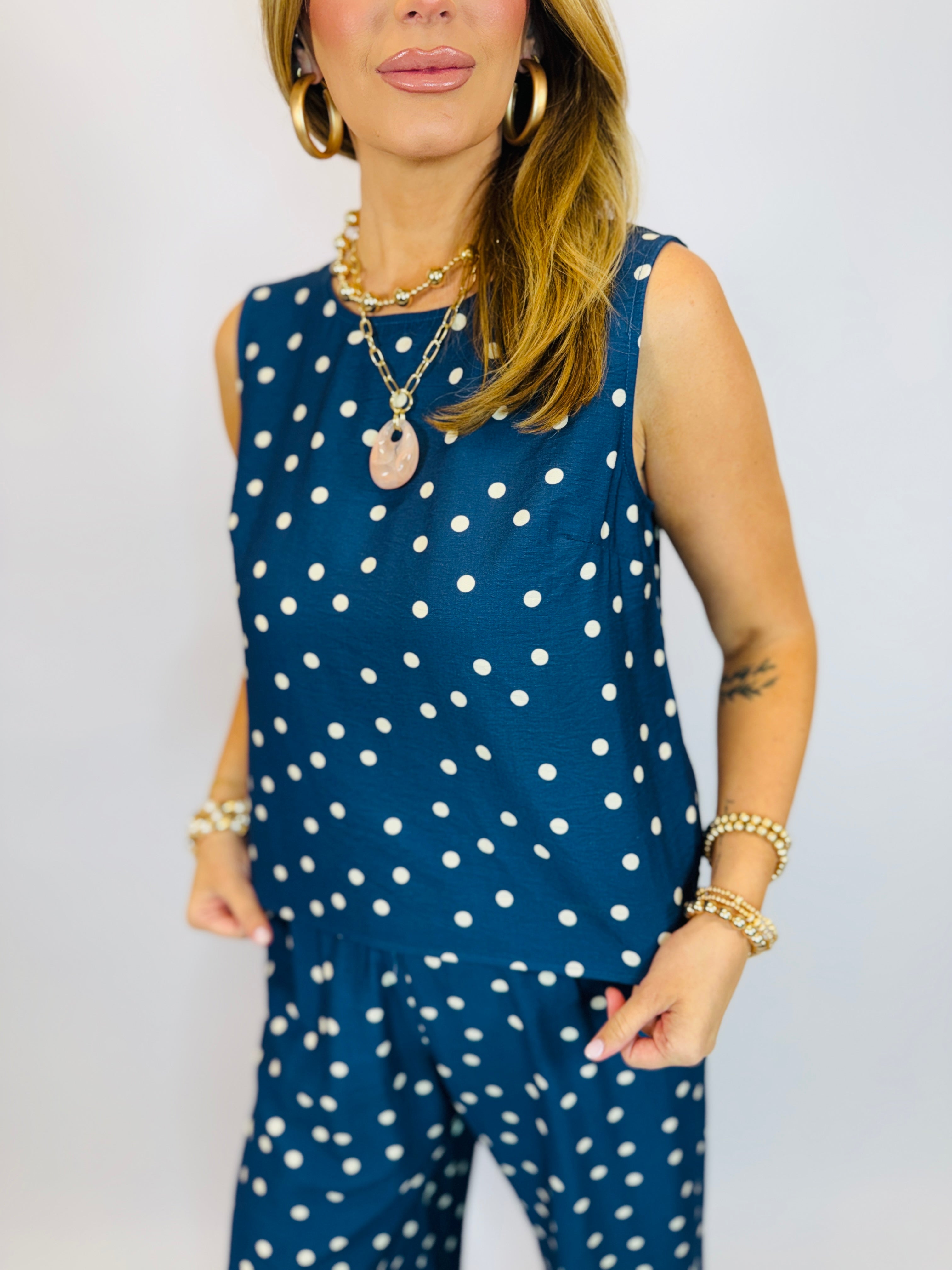 Woman wearing navy blue polka dot sleeveless top with white dots, styled with gold jewelry and bracelets.
