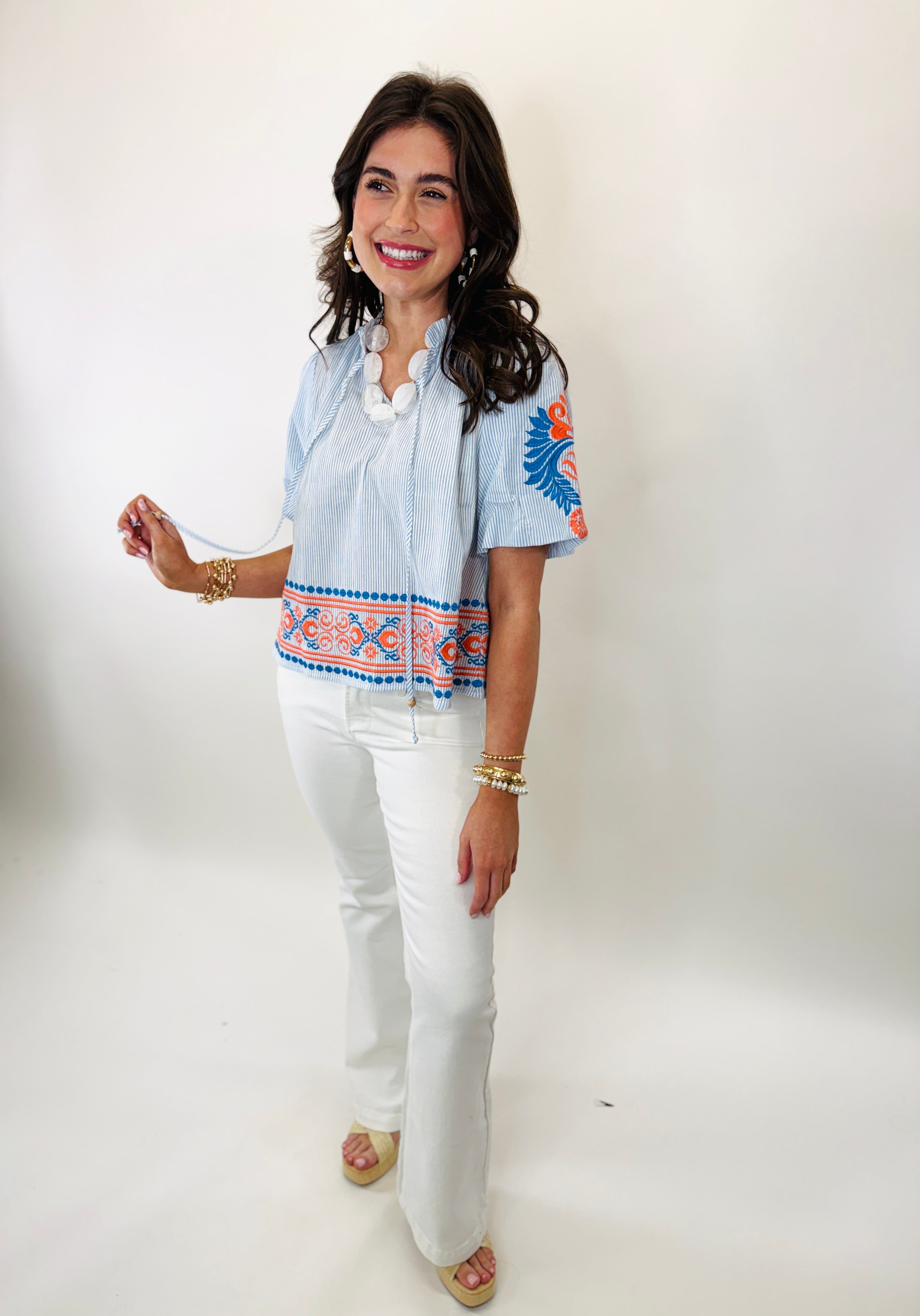 Woman wearing blue striped embroidered Marcie top with white pants and gold jewelry, smiling at camera
