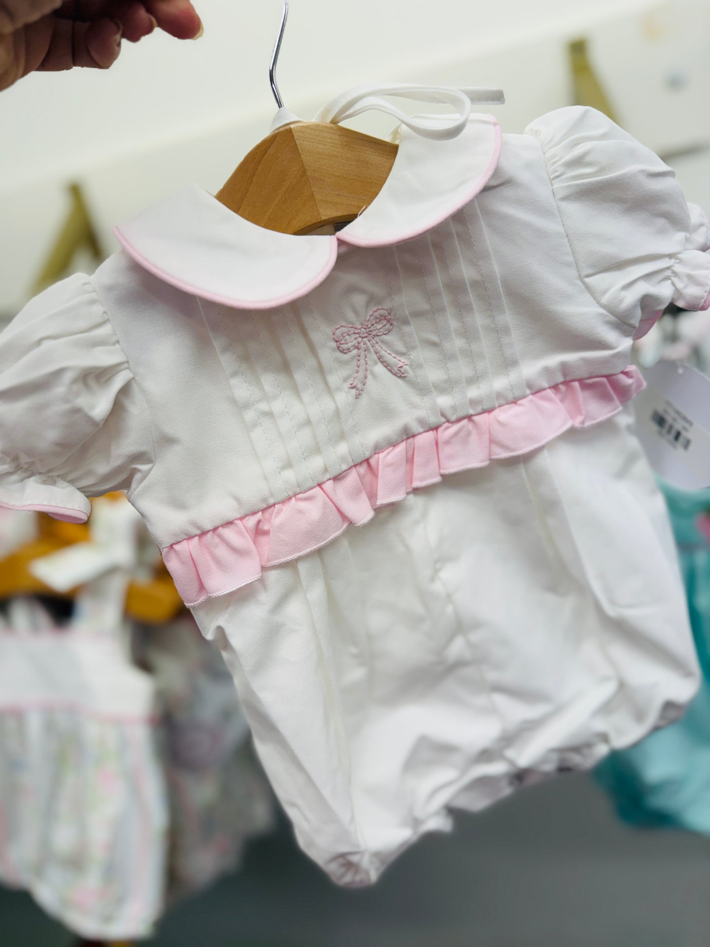 White baby outfit with pink collar trim and embroidered bow detail being held on hanger