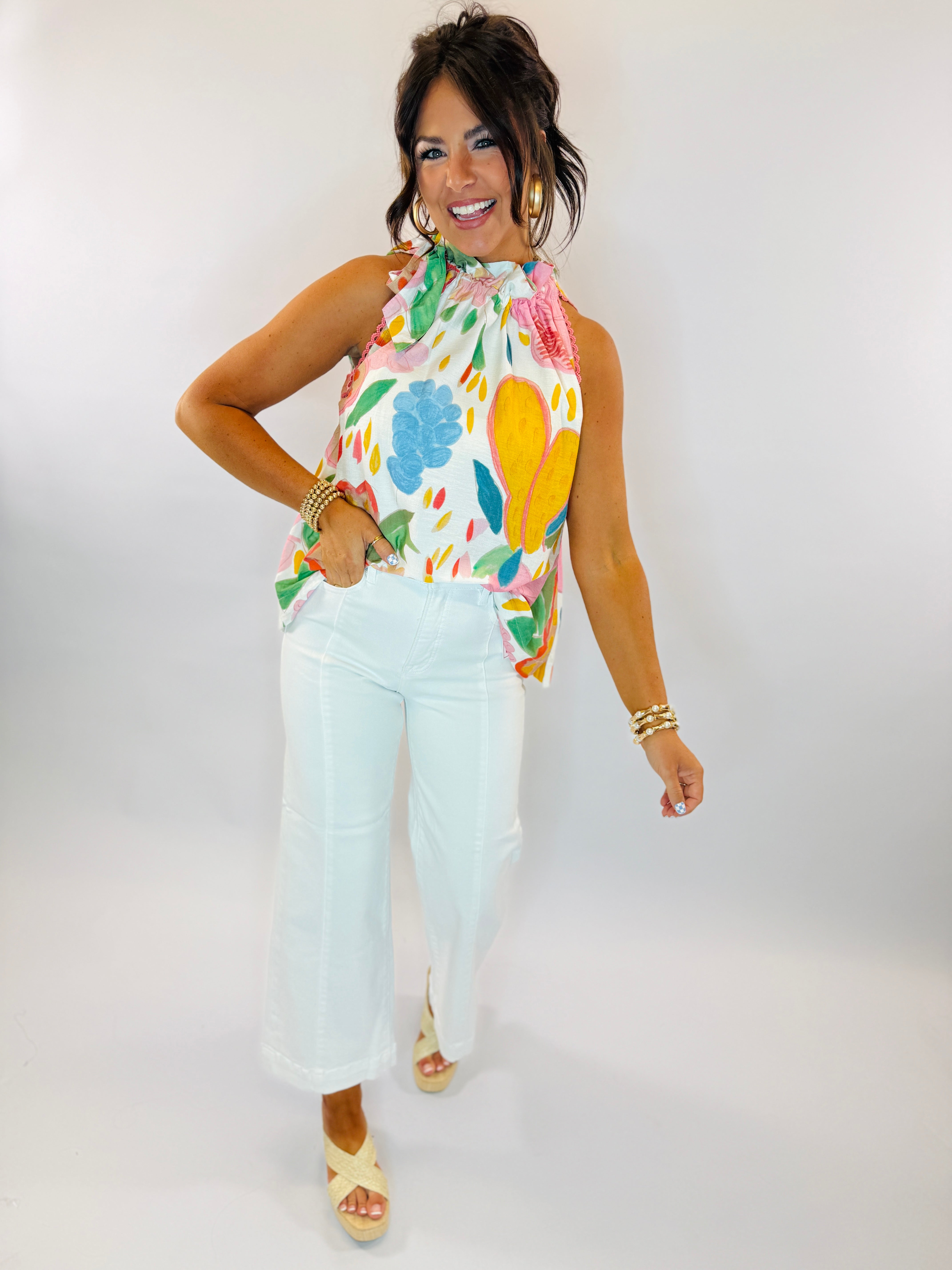 Woman wearing colorful abstract print halter top with high neck and white pants, styled with gold jewelry.
