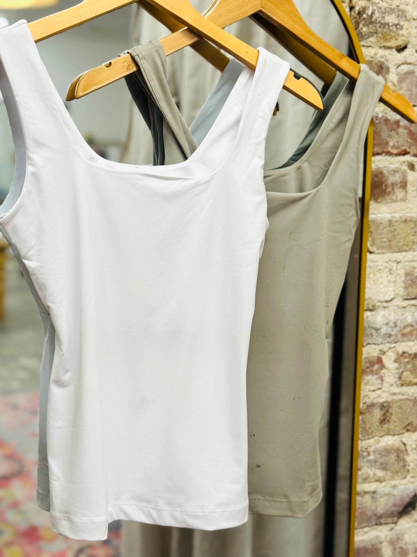 White and sage green Adriana tank tops hanging on wooden hangers against brick wall