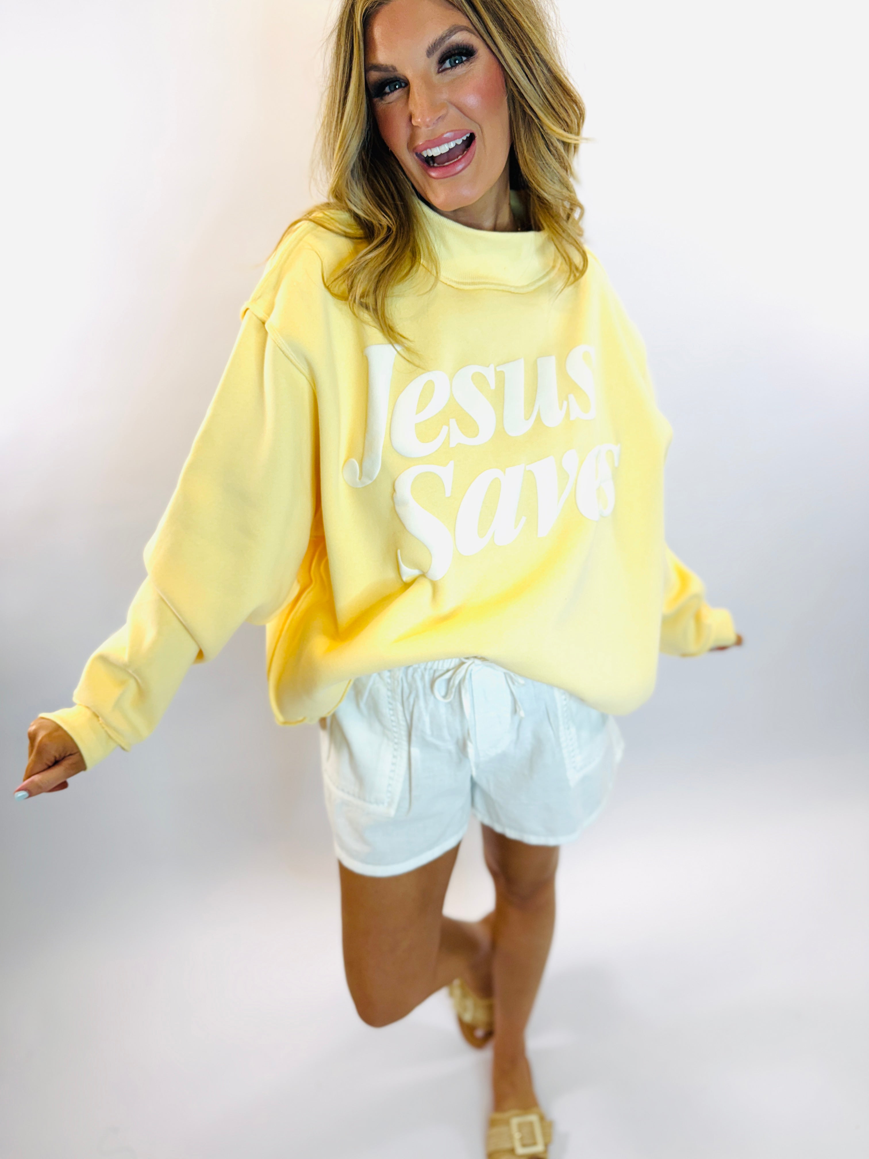 Woman wearing yellow oversized mock neck sweatshirt with "Jesus Saves" text and light blue shorts.
