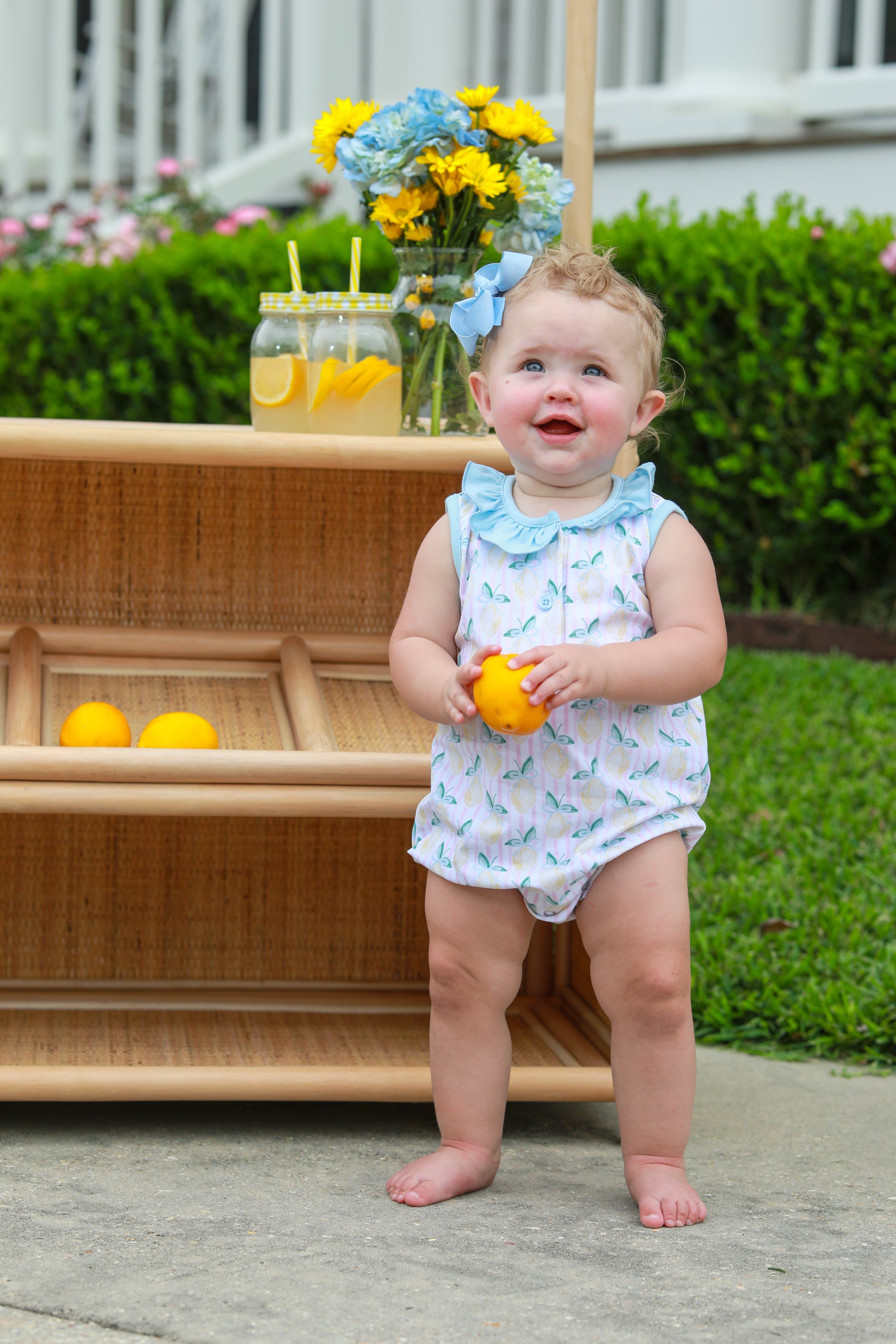 Happy baby in white romper with blue bow headband holding yellow lemon outdoors near lemonade stand