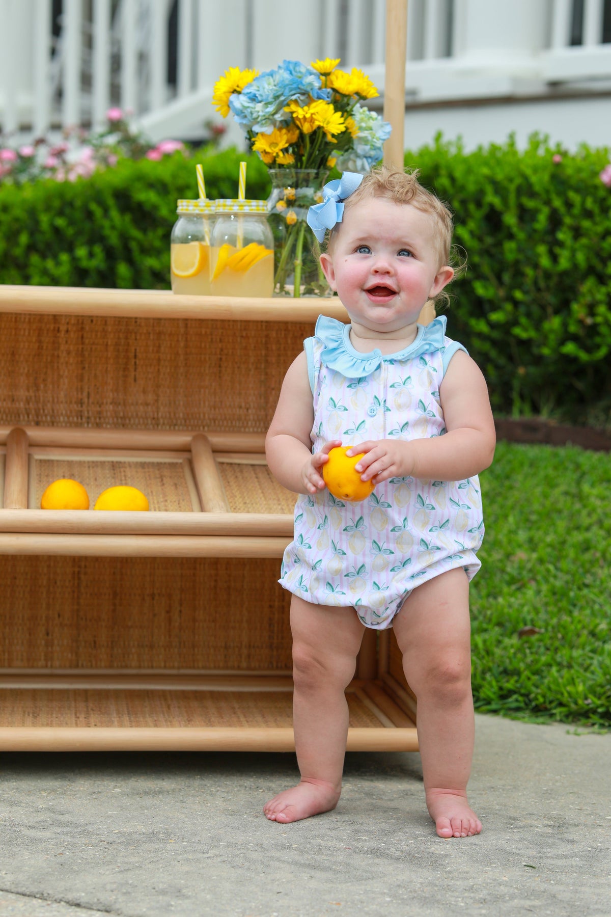 Happy baby in white romper with blue bow headband holding yellow lemon outdoors near lemonade stand