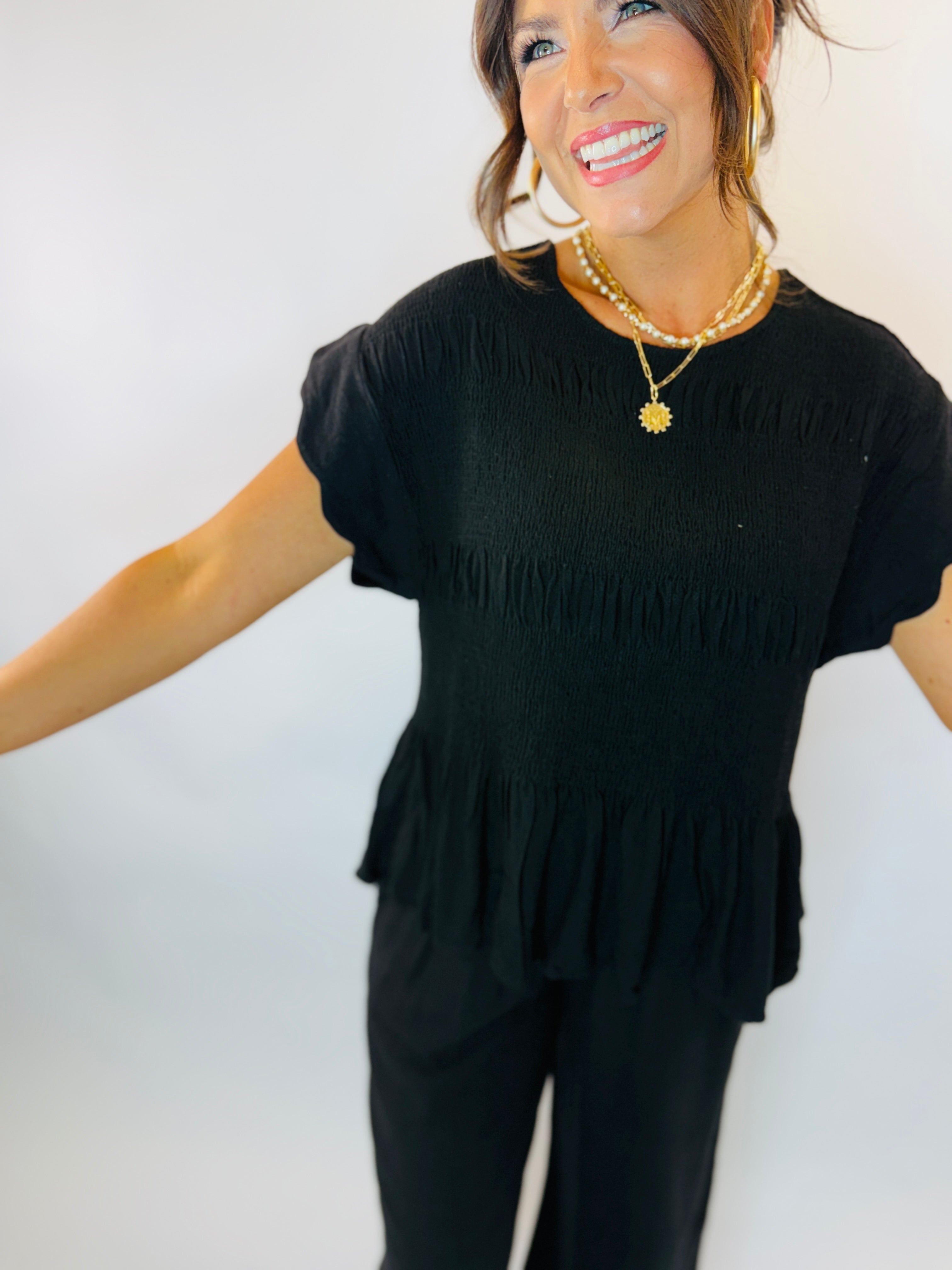 Woman wearing black smocked short-sleeve top with tiered ruffle hem and layered pearl necklaces