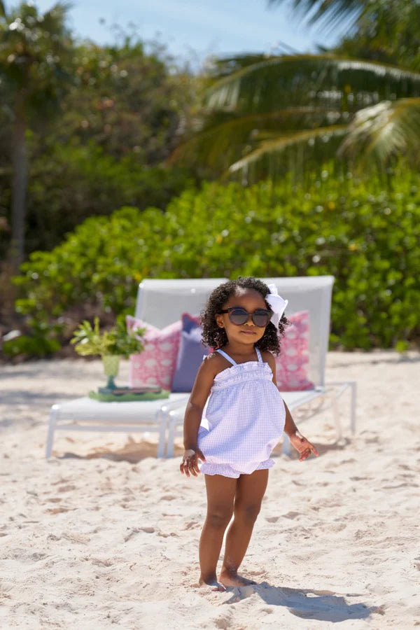 Young child wearing light purple gingham romper with ruffles standing on sandy beach with sunglasses