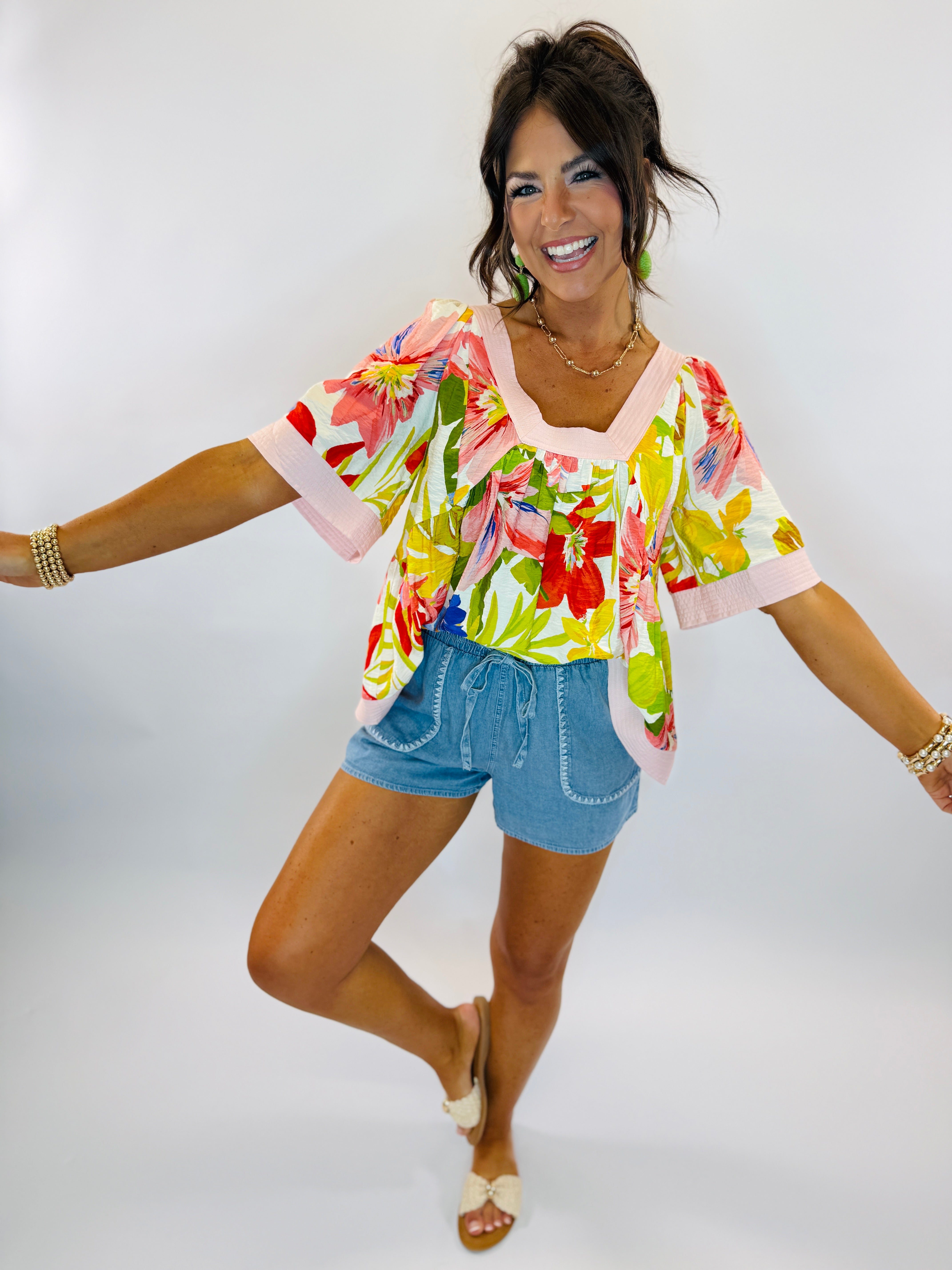 Woman wearing colorful floral Chloe top with pink trim and denim shorts, smiling with arms outstretched