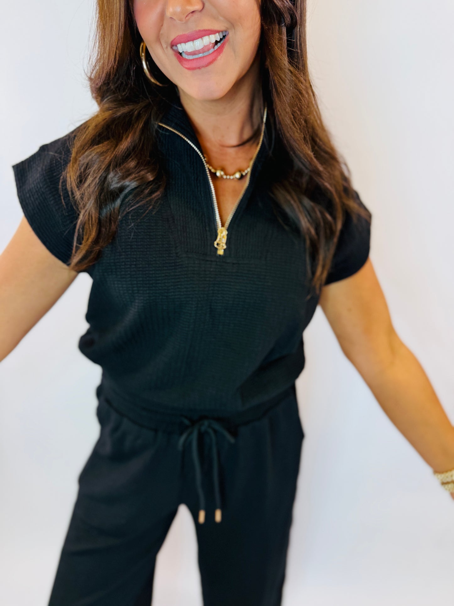 Woman wearing black ribbed short-sleeve zip-up top with drawstring waist and gold jewelry