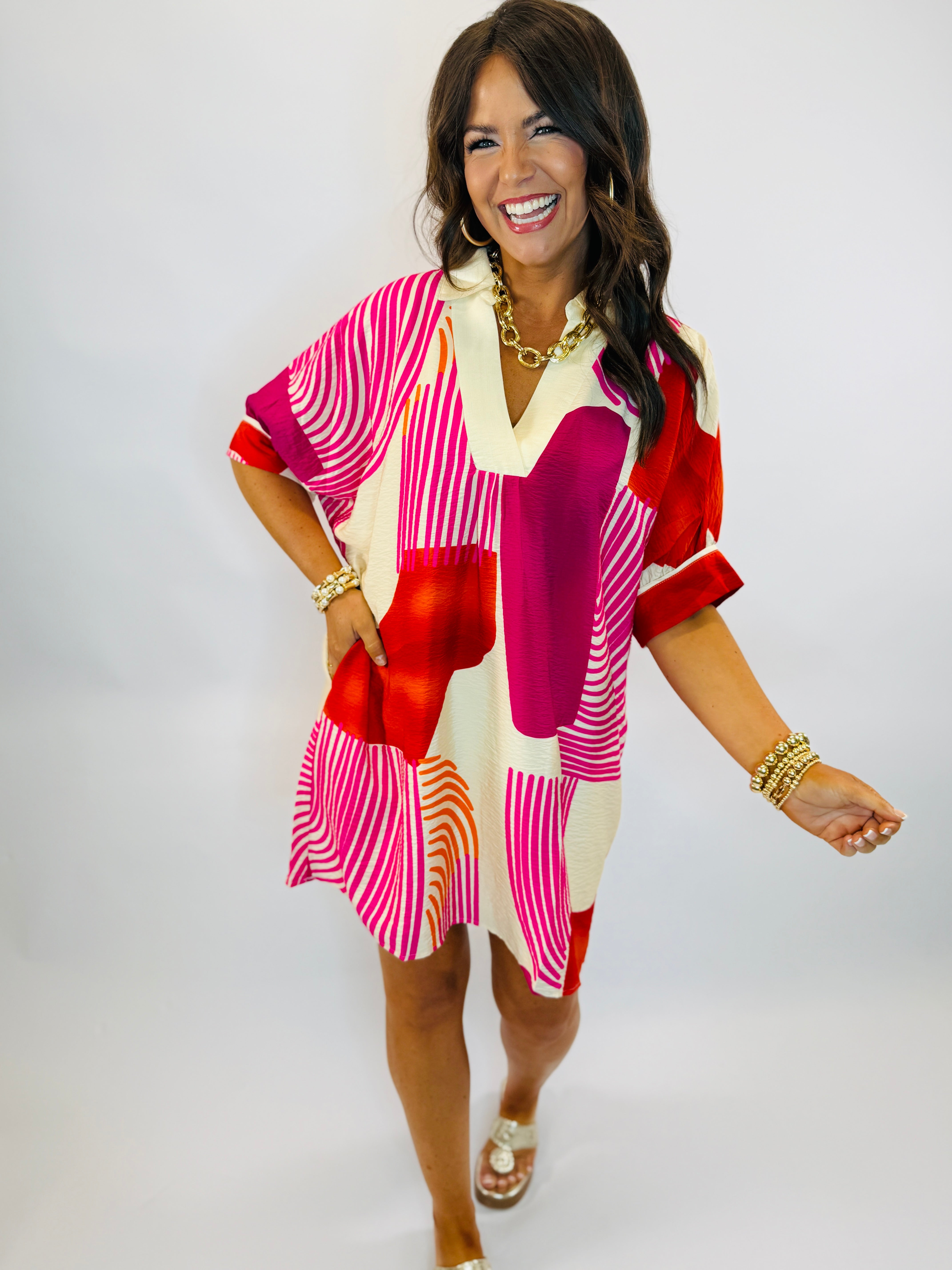 Woman wearing colorful geometric print Hannah dress with pink, red, and white patterns, styled with gold jewelry and sandals.
