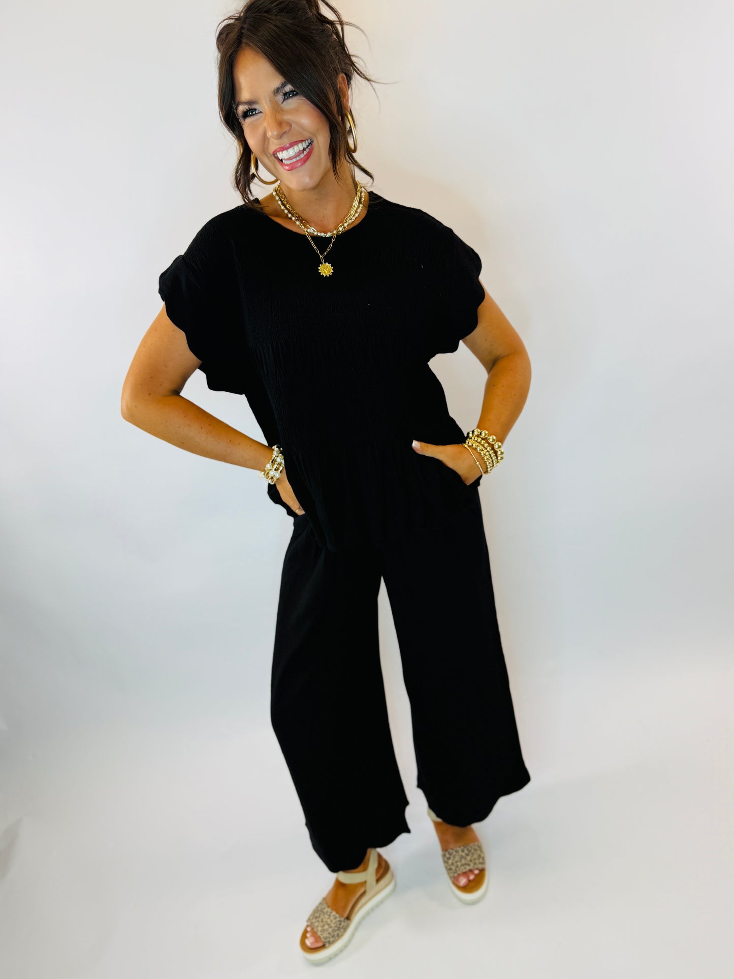Woman wearing black short-sleeve top with ruffle details and gold jewelry, smiling against white background.