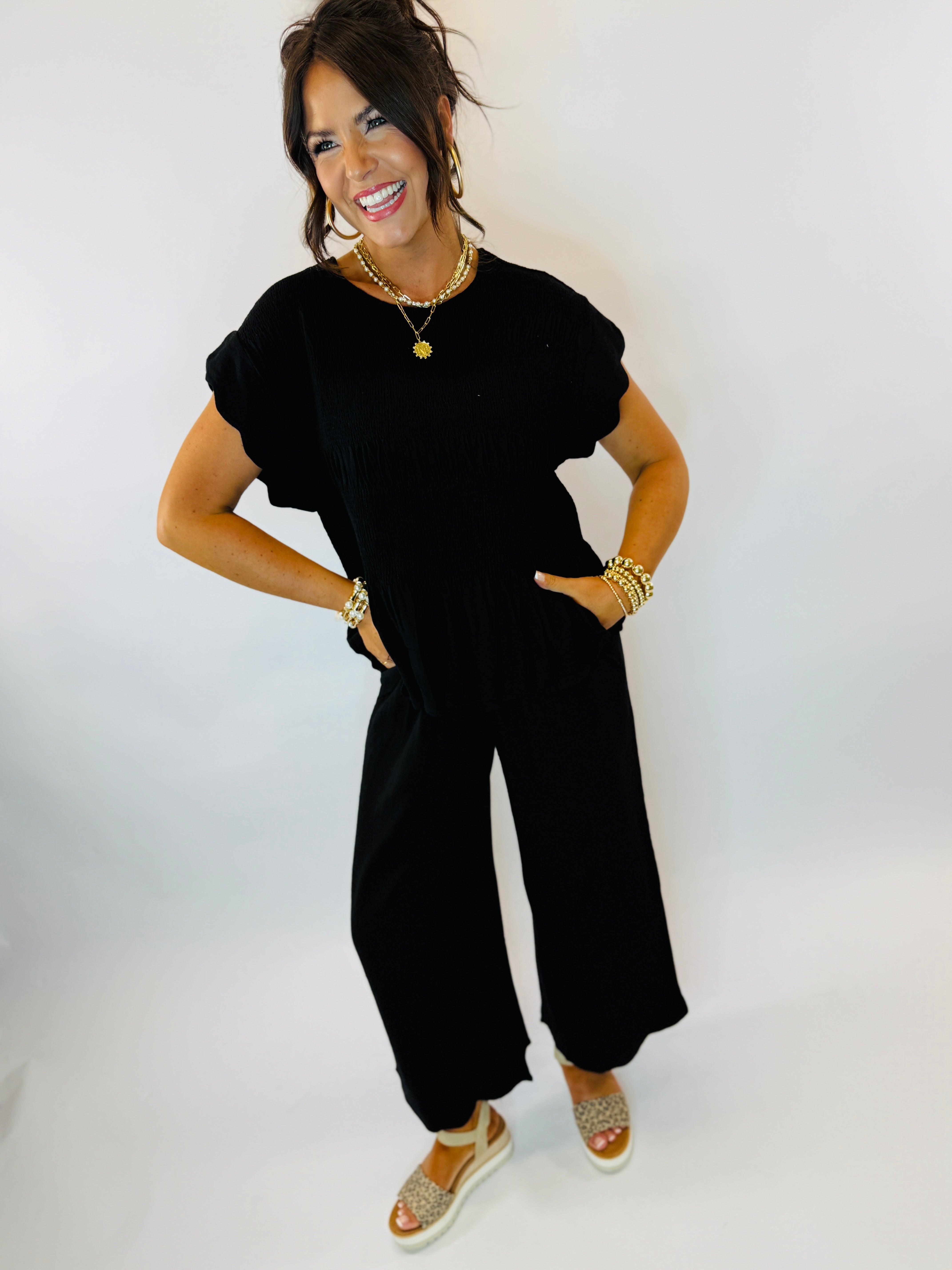 Woman wearing black short-sleeve top with ruffle details and gold jewelry, smiling against white background.