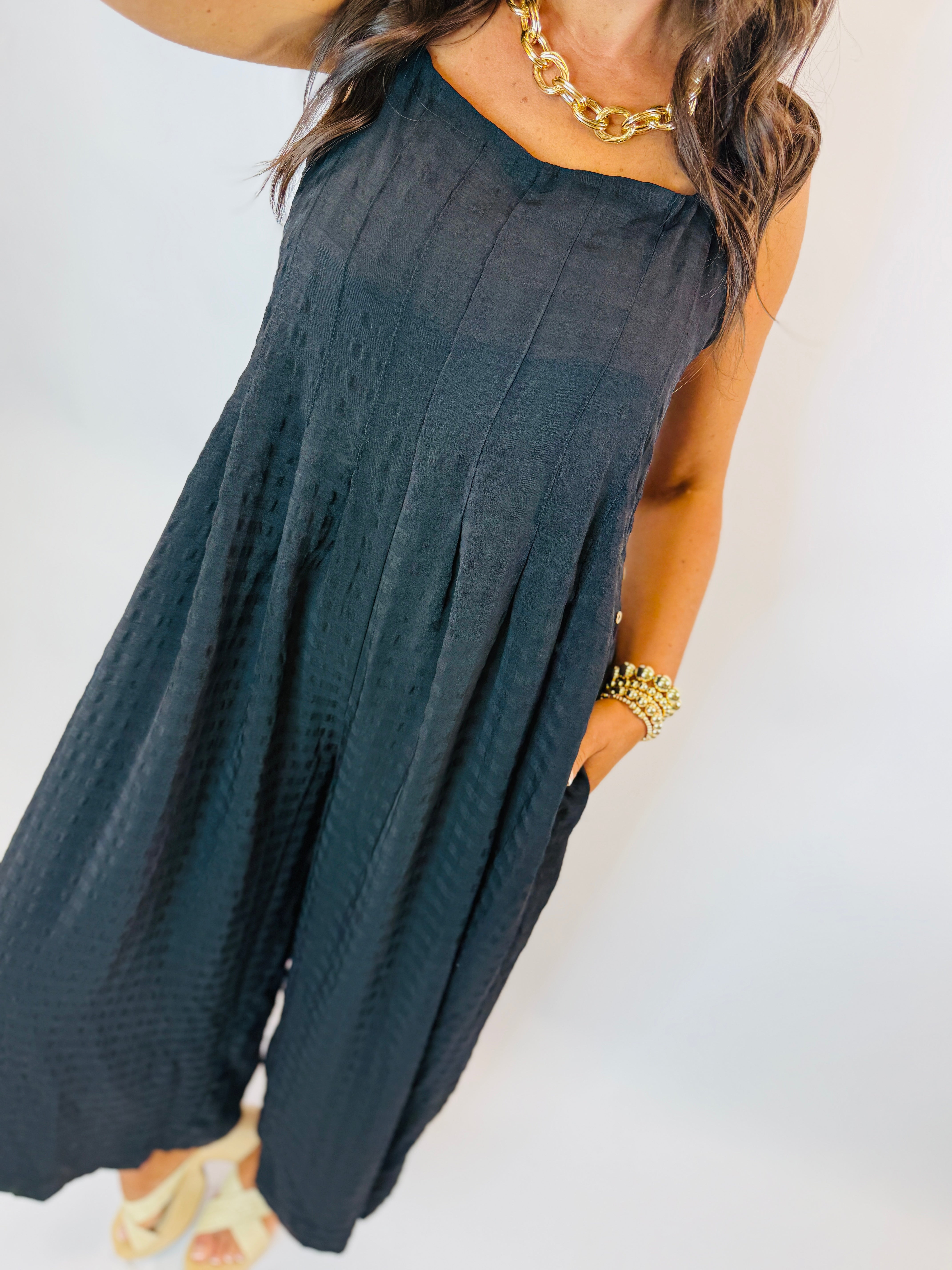 Woman wearing navy blue sleeveless pleated jumpsuit with gold chain necklace and bracelet accessories.