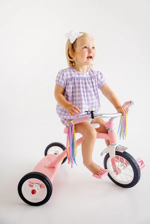 Young girl in purple gingham dress riding pink tricycle with colorful streamers on handlebars