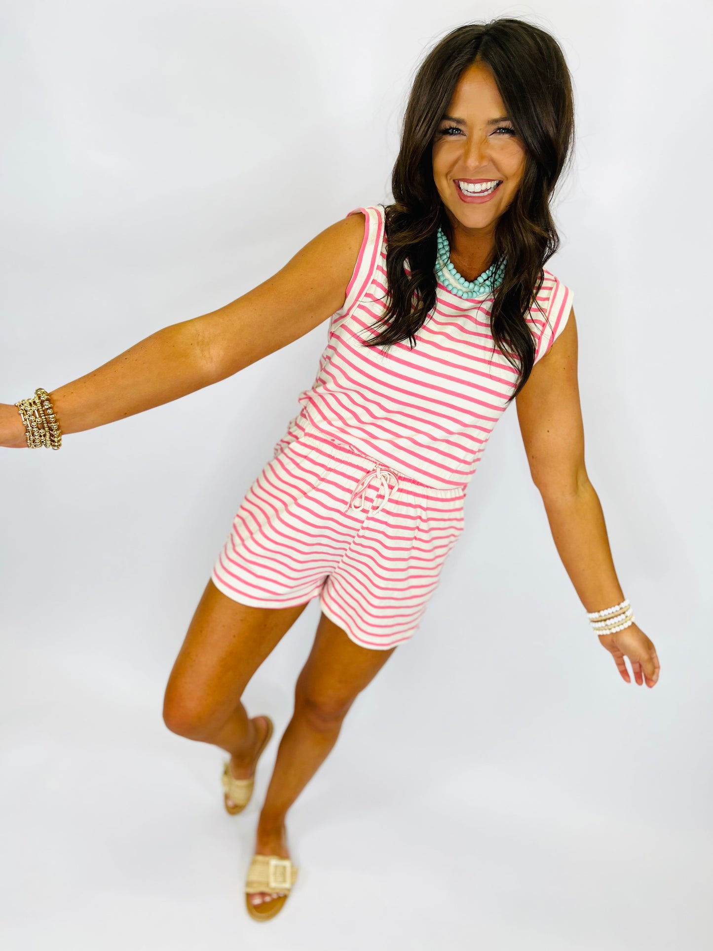 Woman wearing pink and white striped sleeveless romper with drawstring waist, accessorized with turquoise necklace and gol...