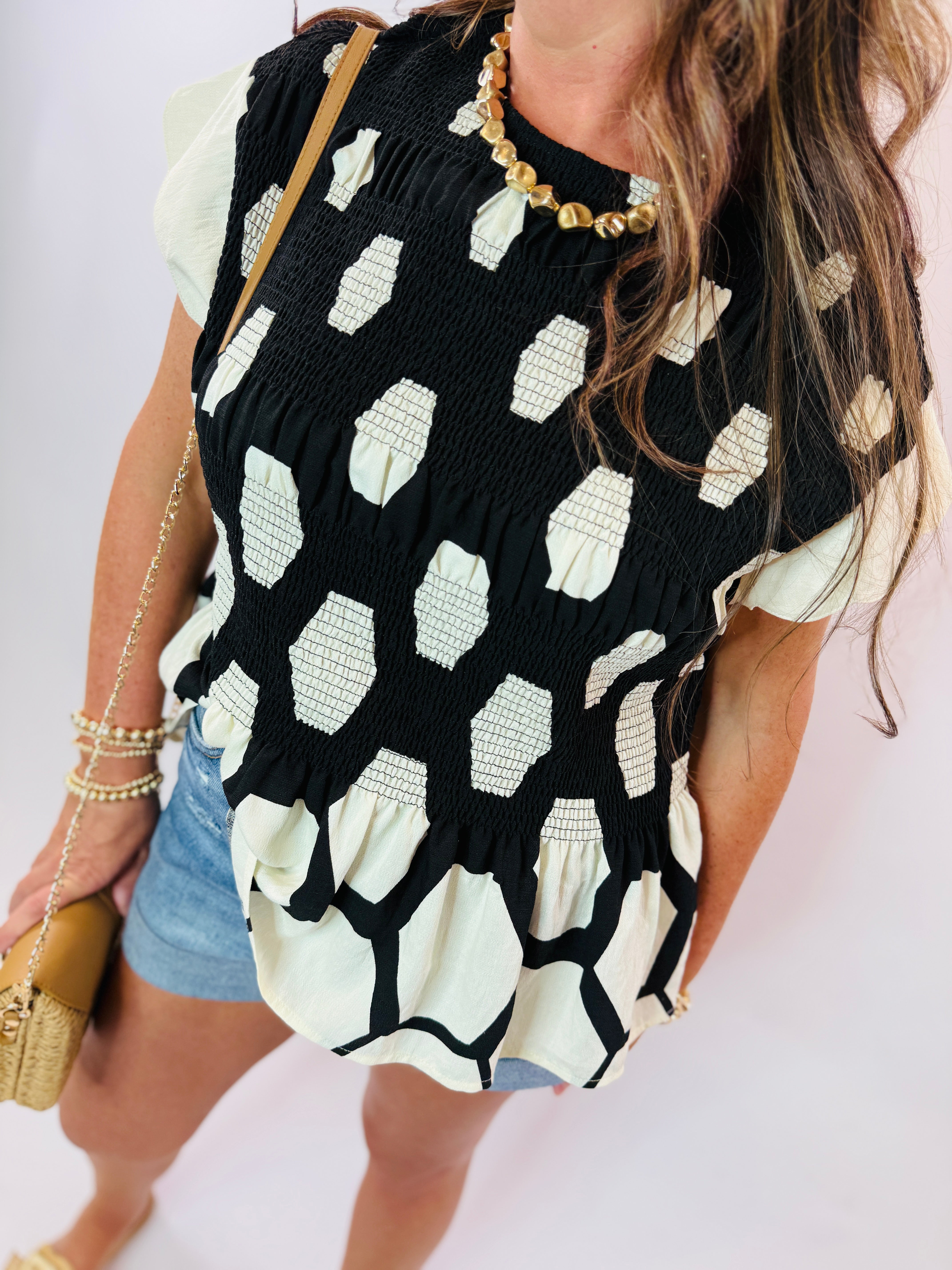 Black and white polka dot smocked top with short sleeves worn with denim shorts and gold jewelry.