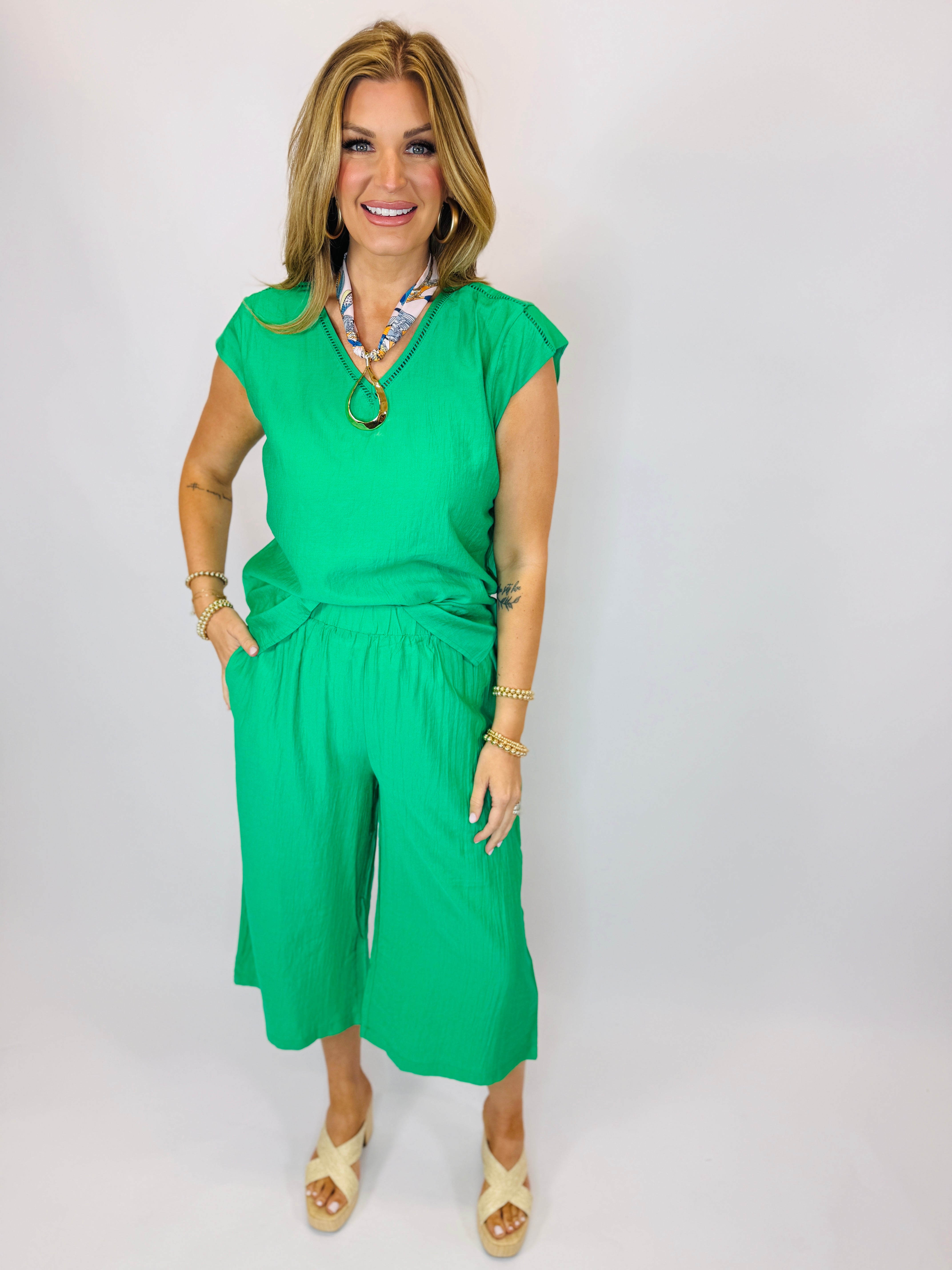 Woman wearing bright green short-sleeve top and matching wide-leg cropped pants with gold jewelry and cream sandals.
