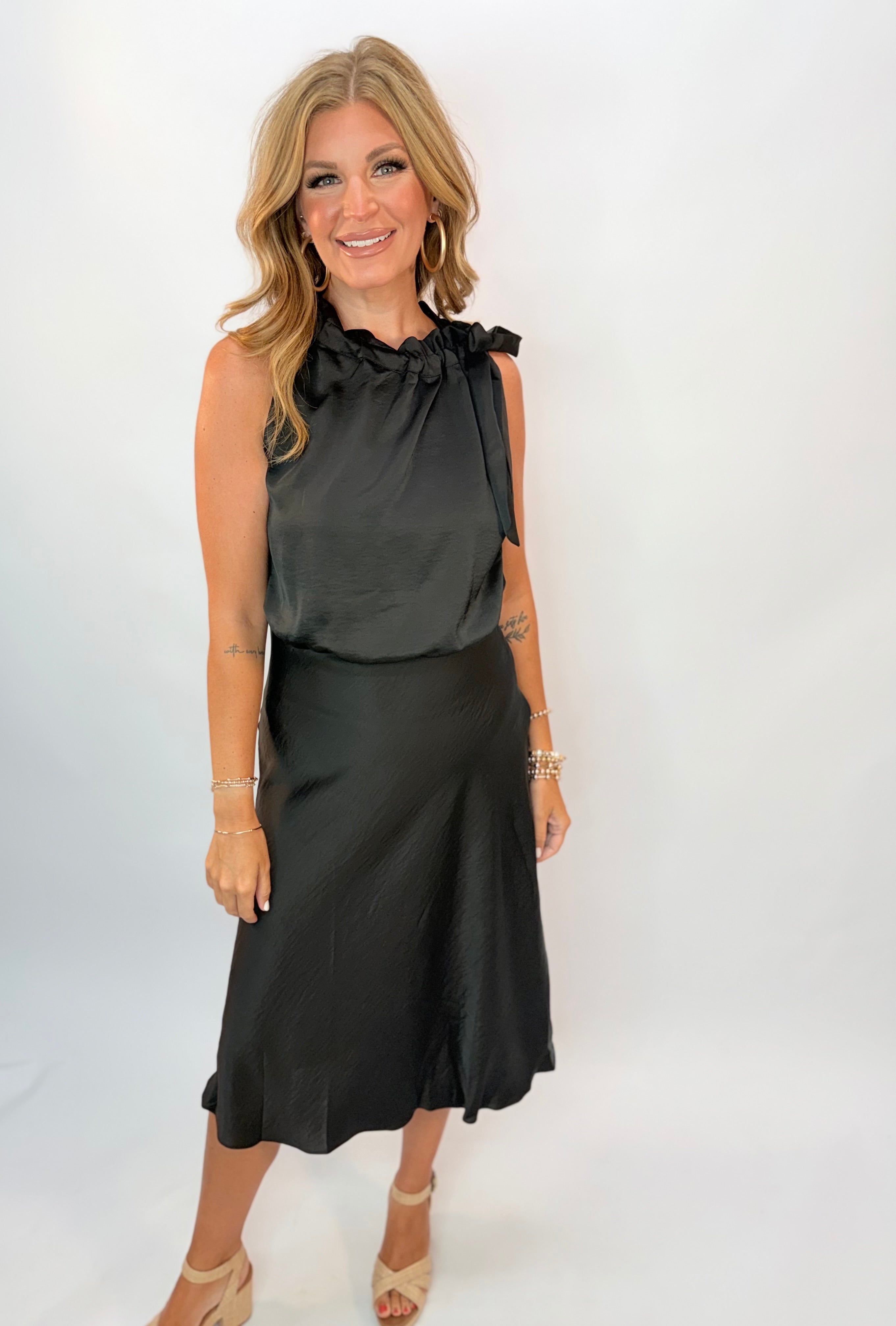 Black satin midi dress with ruched halter neckline and side tie detail, styled with nude heels.