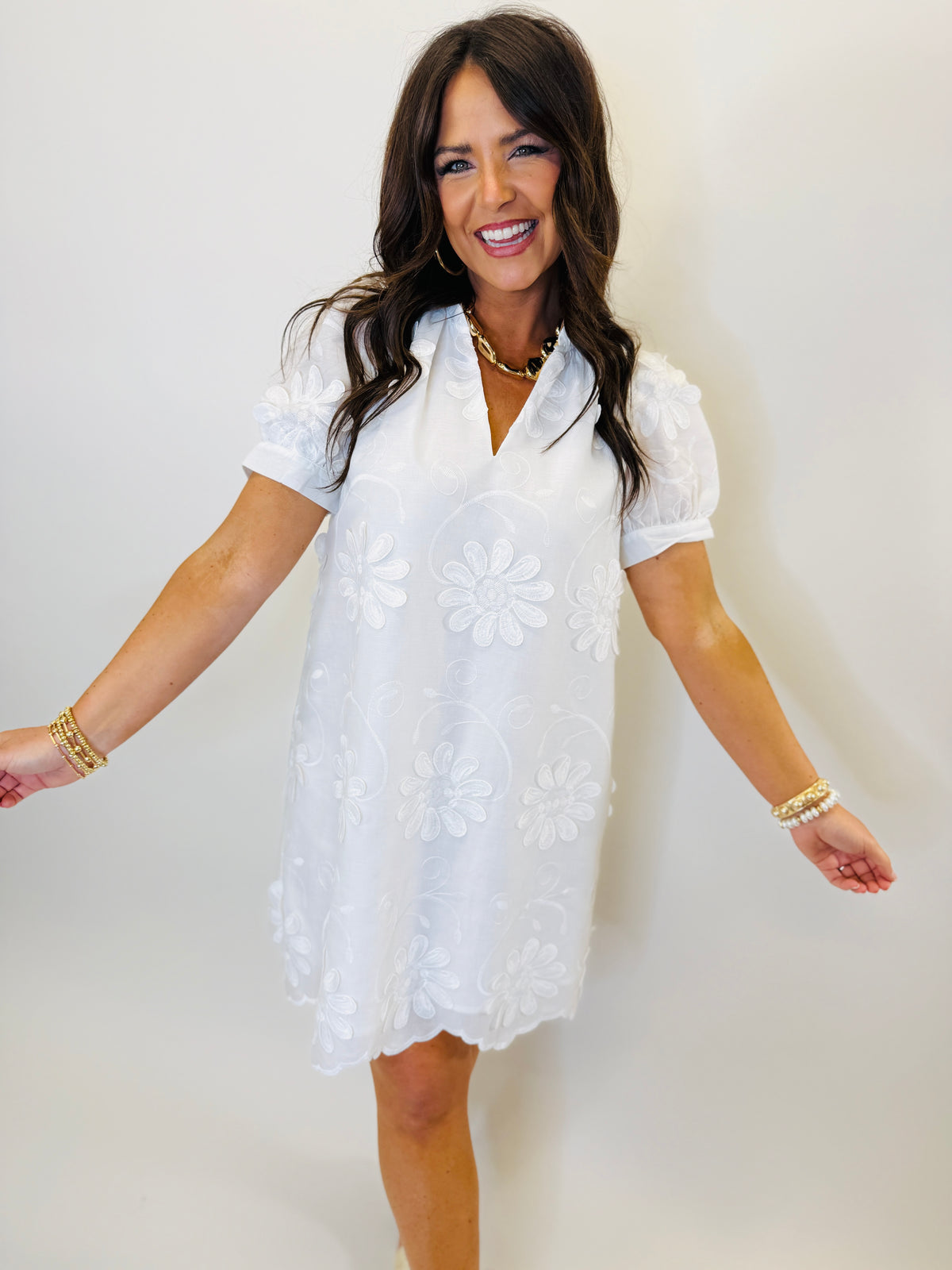Woman wearing white short-sleeve embroidered dress with floral pattern and gold jewelry, smiling with arms outstretched.