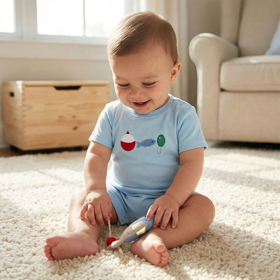 Baby boy in light blue fishing-themed romper sitting on carpet playing with wooden toy
