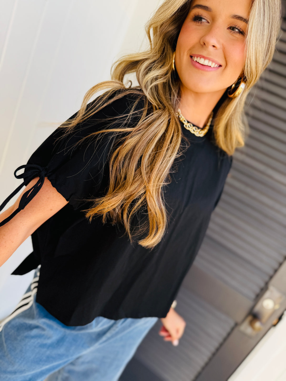 Woman wearing black Juliana top with tie sleeves and chain necklace, smiling while sitting