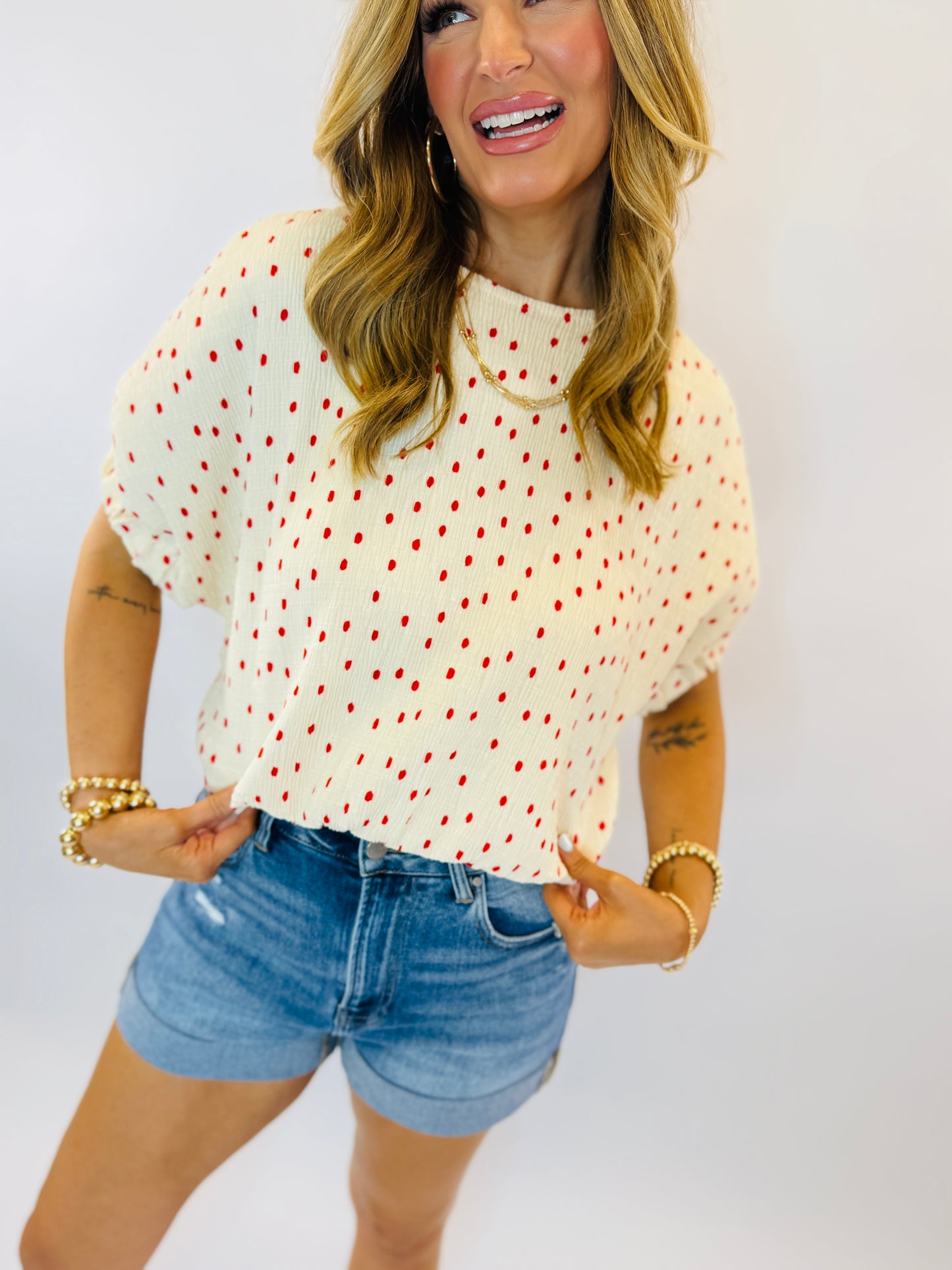 Woman wearing cream knit sweater with red polka dots and denim shorts, smiling at camera