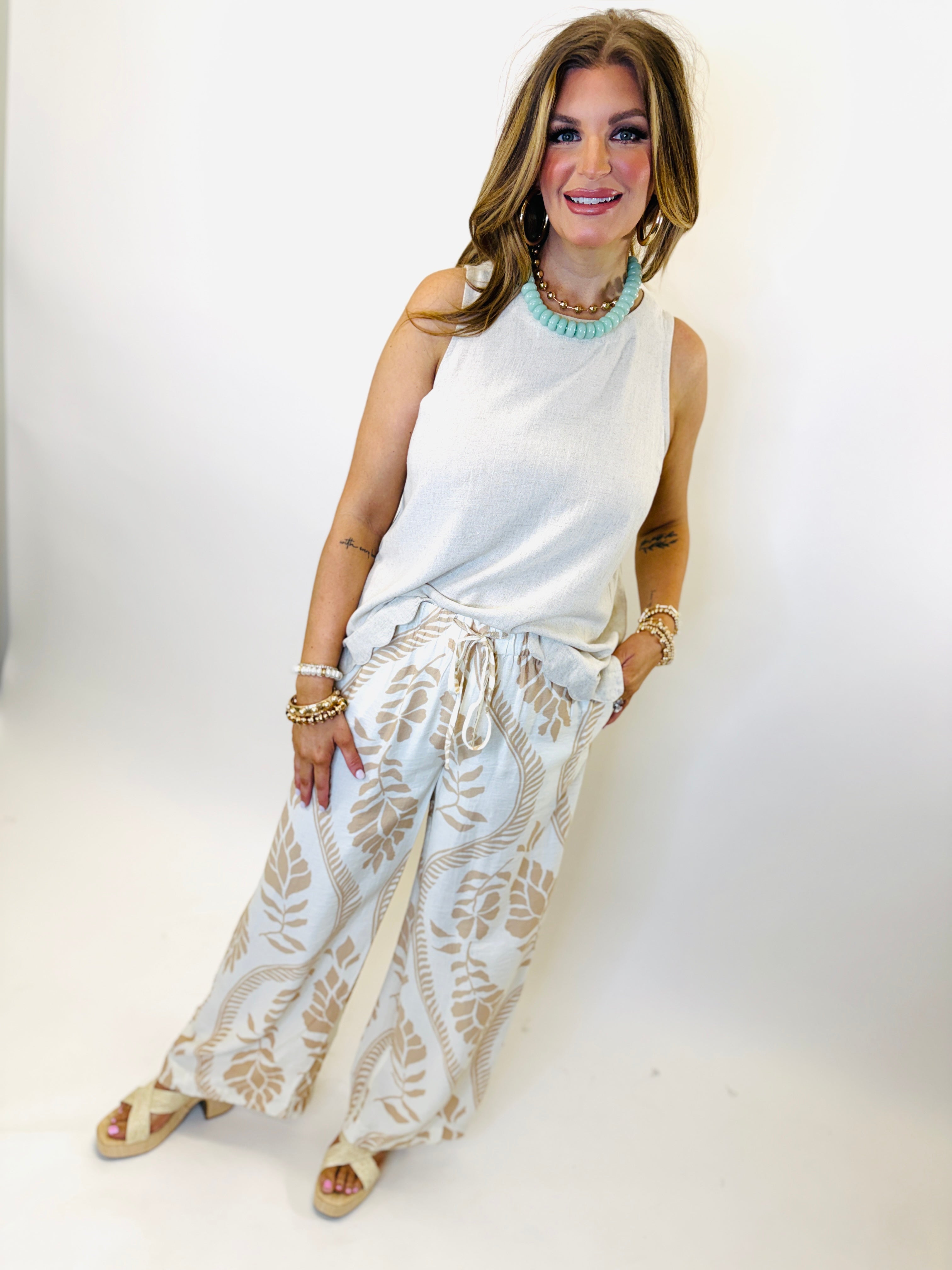 Woman wearing white sleeveless Kenslee top with floral print pants, turquoise necklace, and espadrille sandals