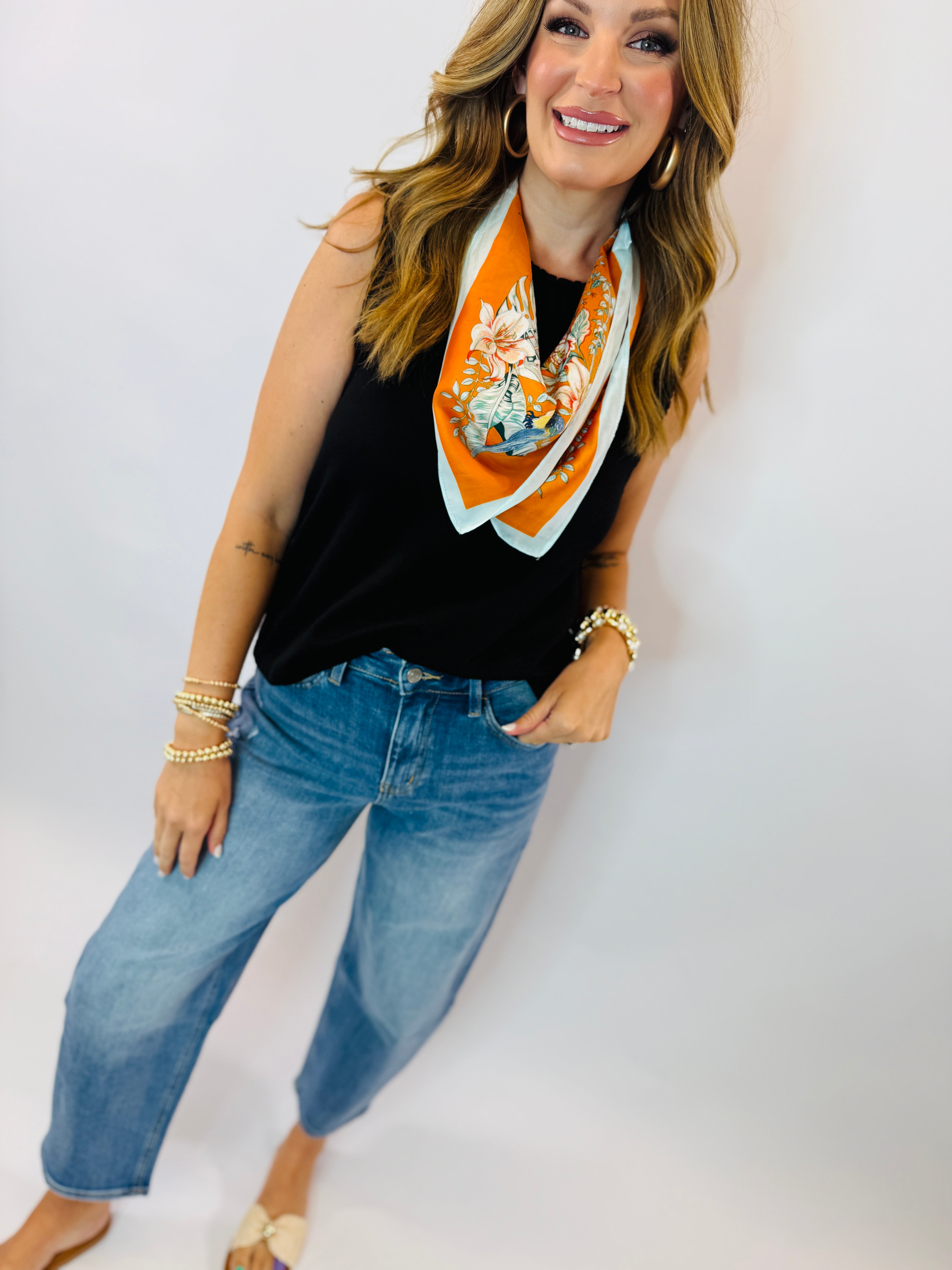 Woman wearing black sleeveless Georgie top with orange floral scarf, blue jeans, and gold jewelry accessories.