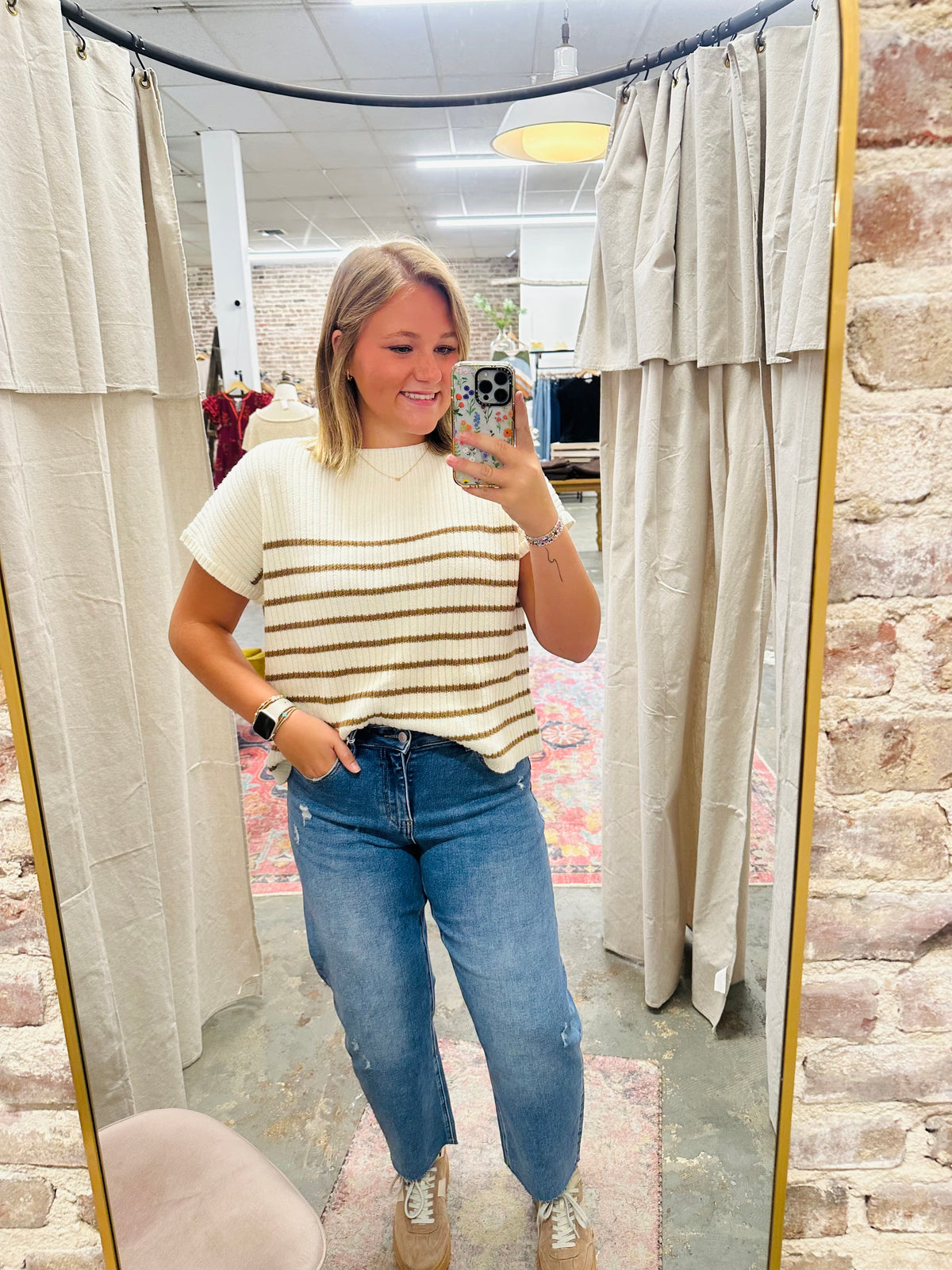 Woman wearing distressed blue jeans and striped sweater taking mirror selfie in store fitting room