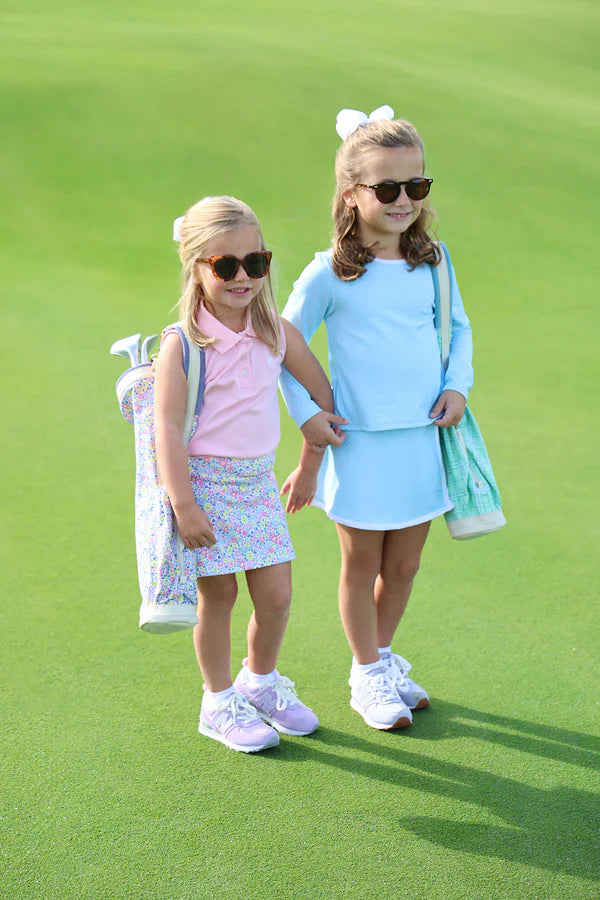 Two young girls wearing sunglasses and colorful skorts standing on green grass holding golf equipment.
