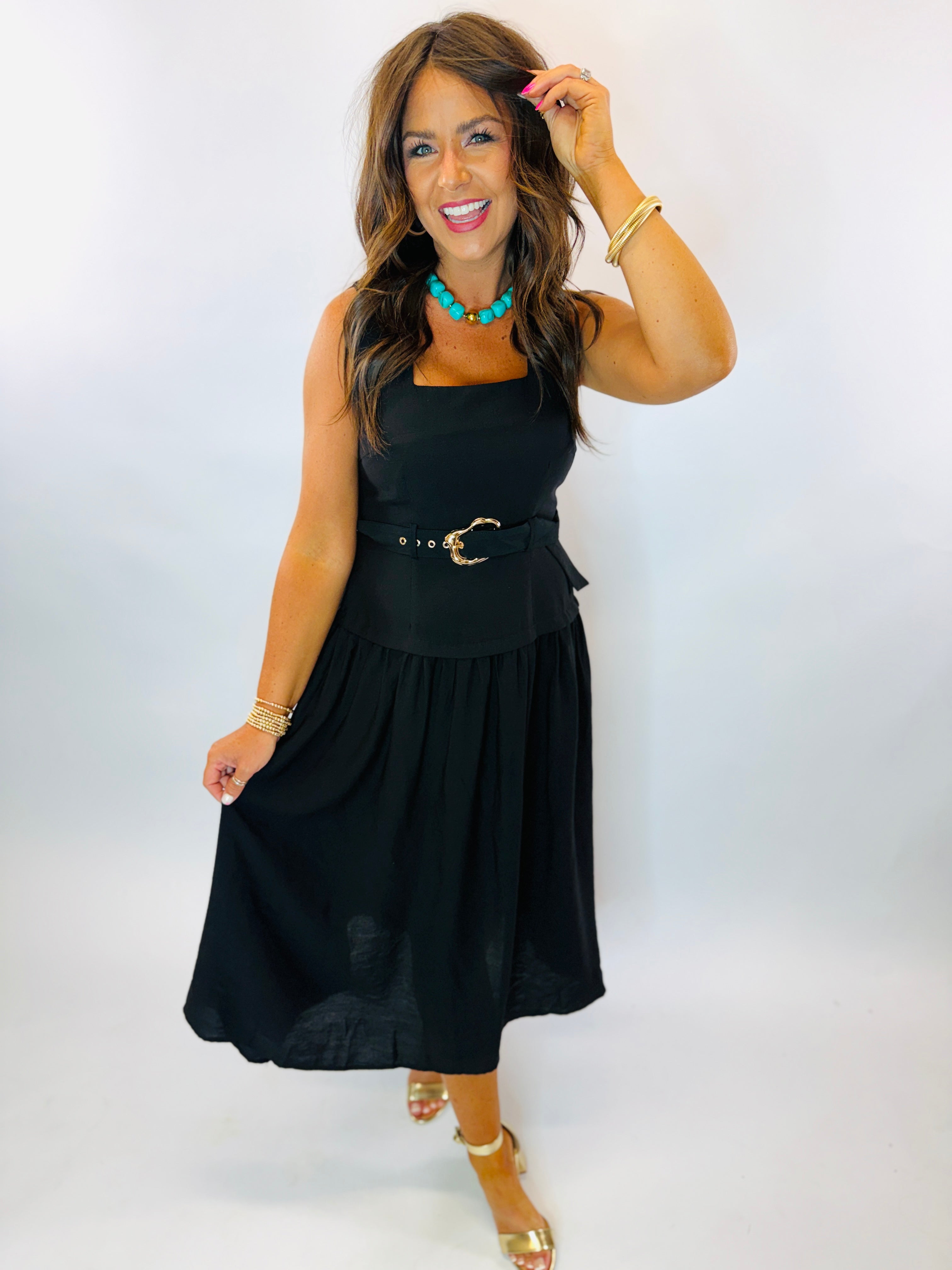 Woman wearing black sleeveless midi dress with gold belt buckle, styled with turquoise necklace and gold sandals.