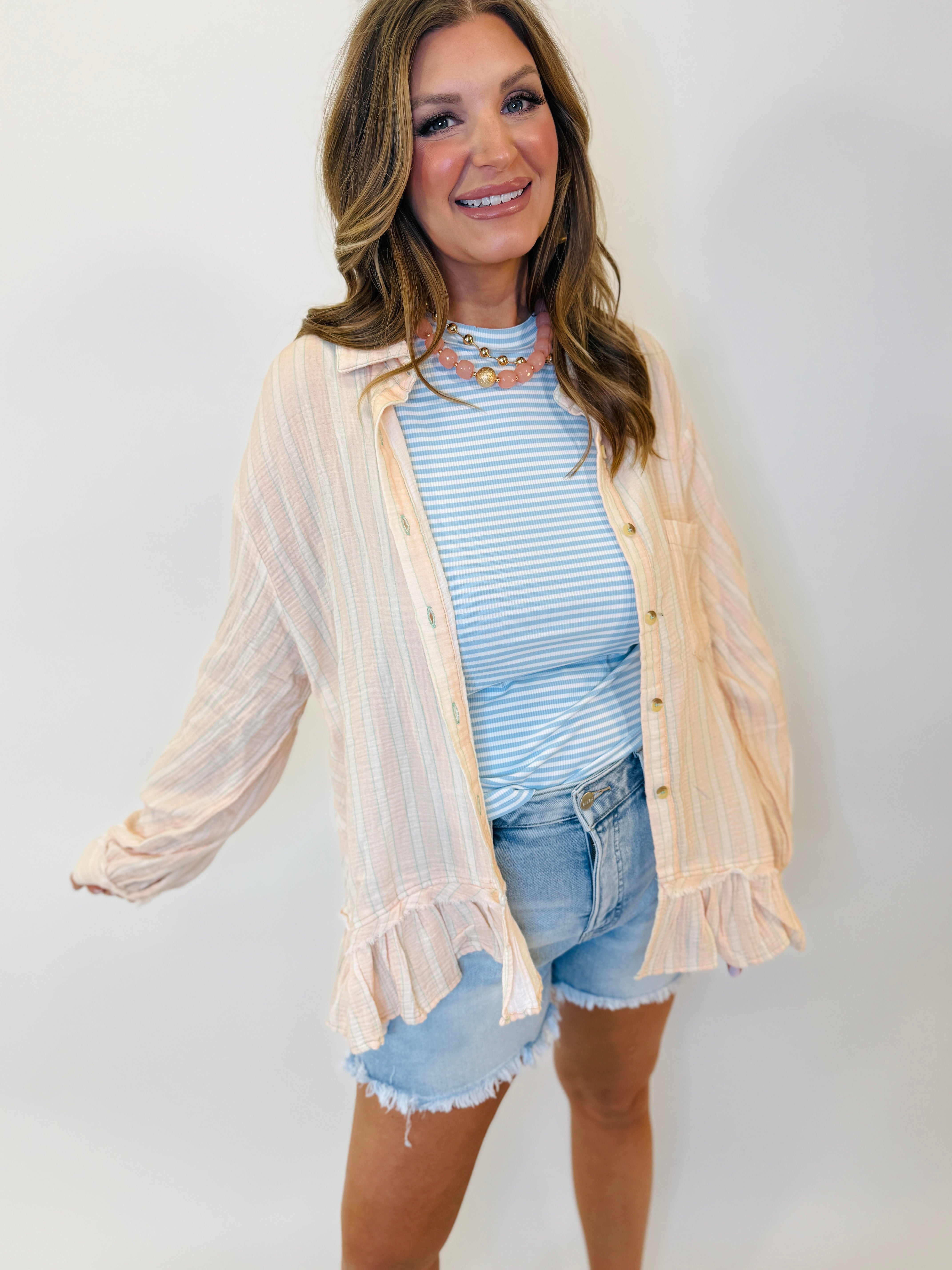 Woman wearing cream ribbed button-up cardigan with ruffle trim over blue striped top and denim shorts