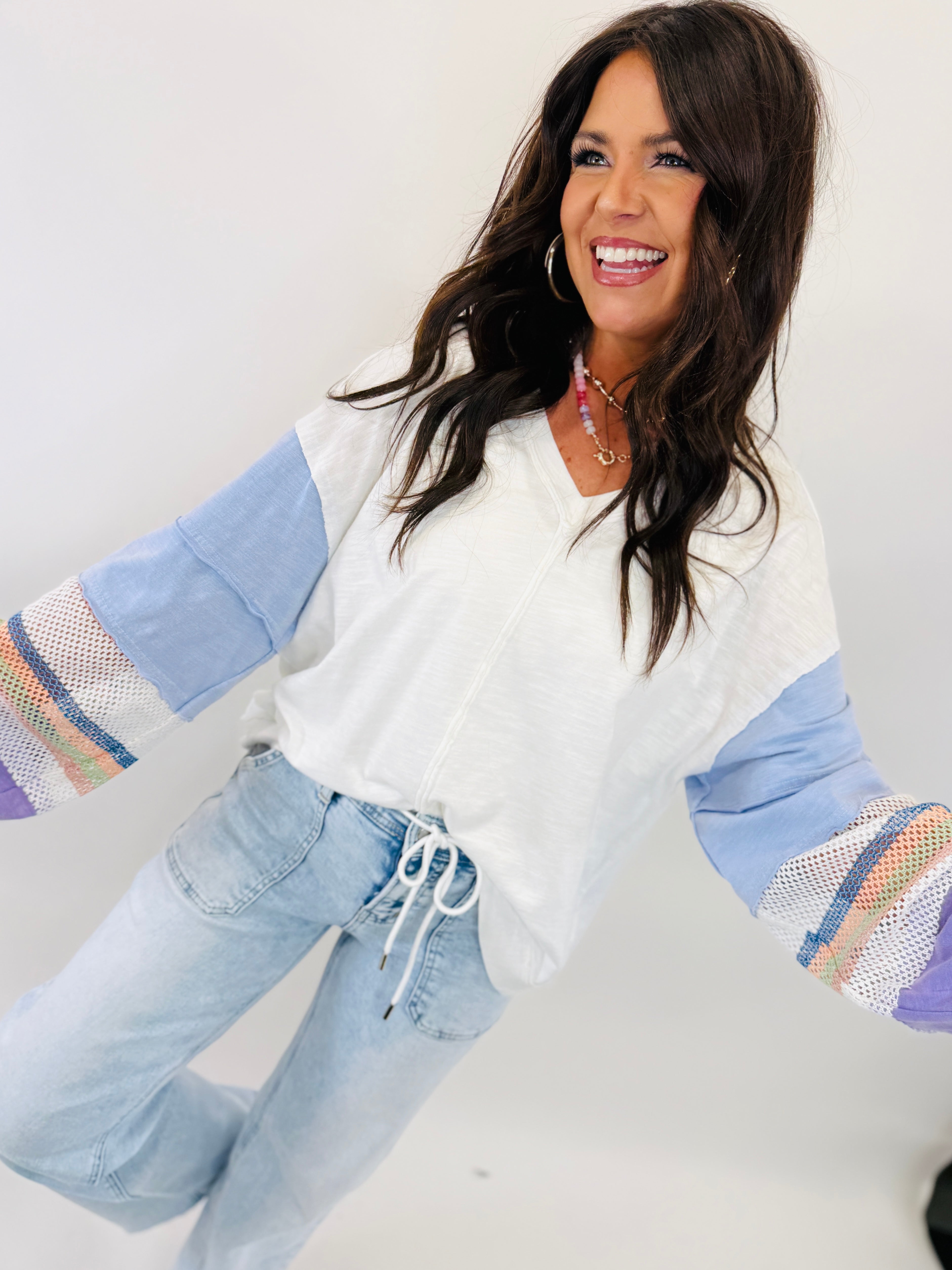 Woman wearing white long-sleeve top with blue sleeves and colorful striped cuffs, paired with light blue jeans.