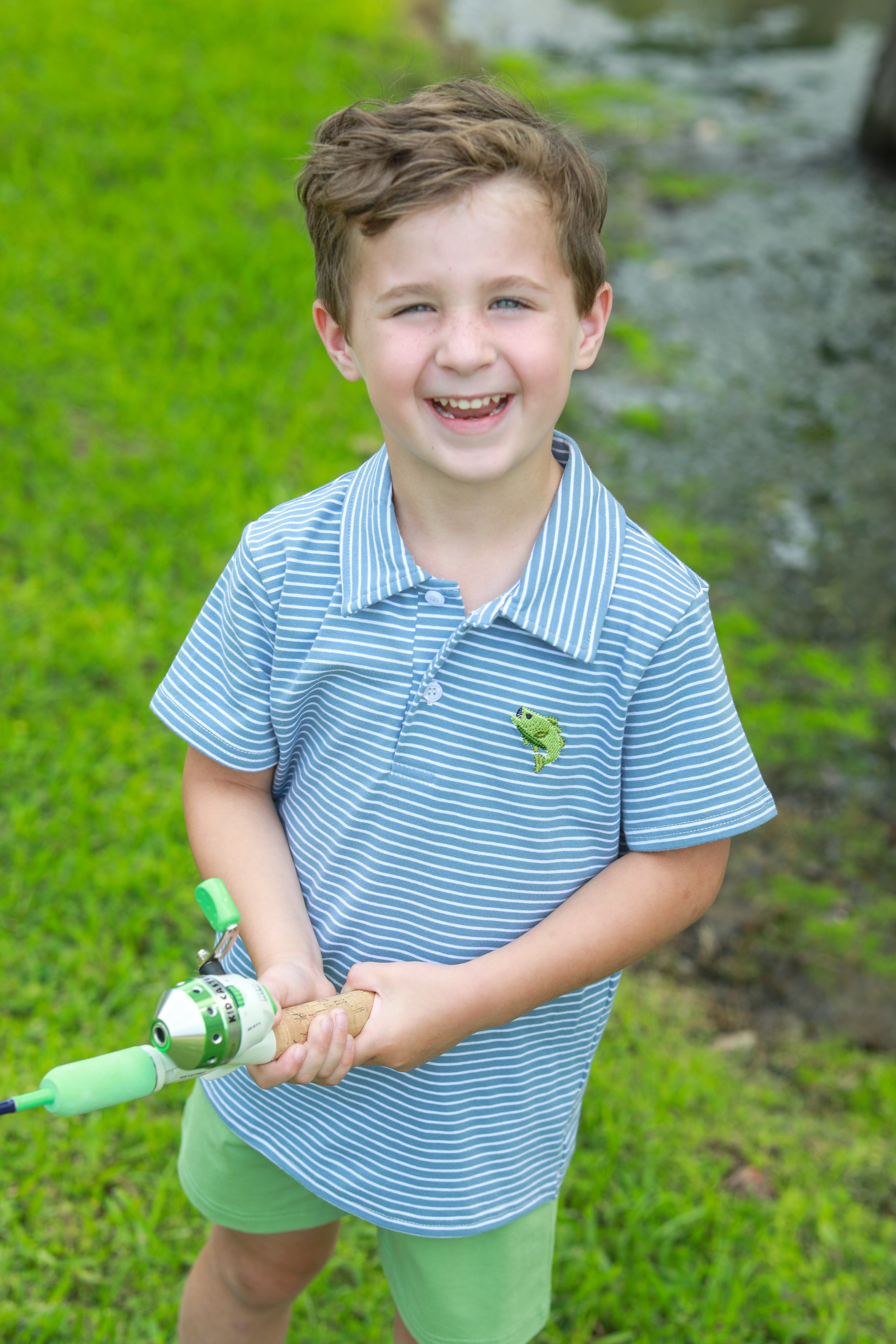 Young boy wearing blue and white striped polo shirt with fish emblem, holding green fishing rod and smiling outdoors.