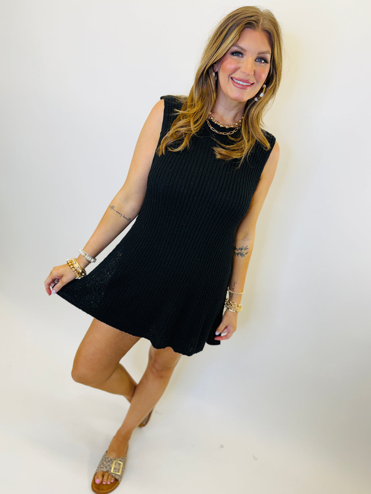 Woman wearing black ribbed sleeveless mini dress with gold jewelry and sandals against white background