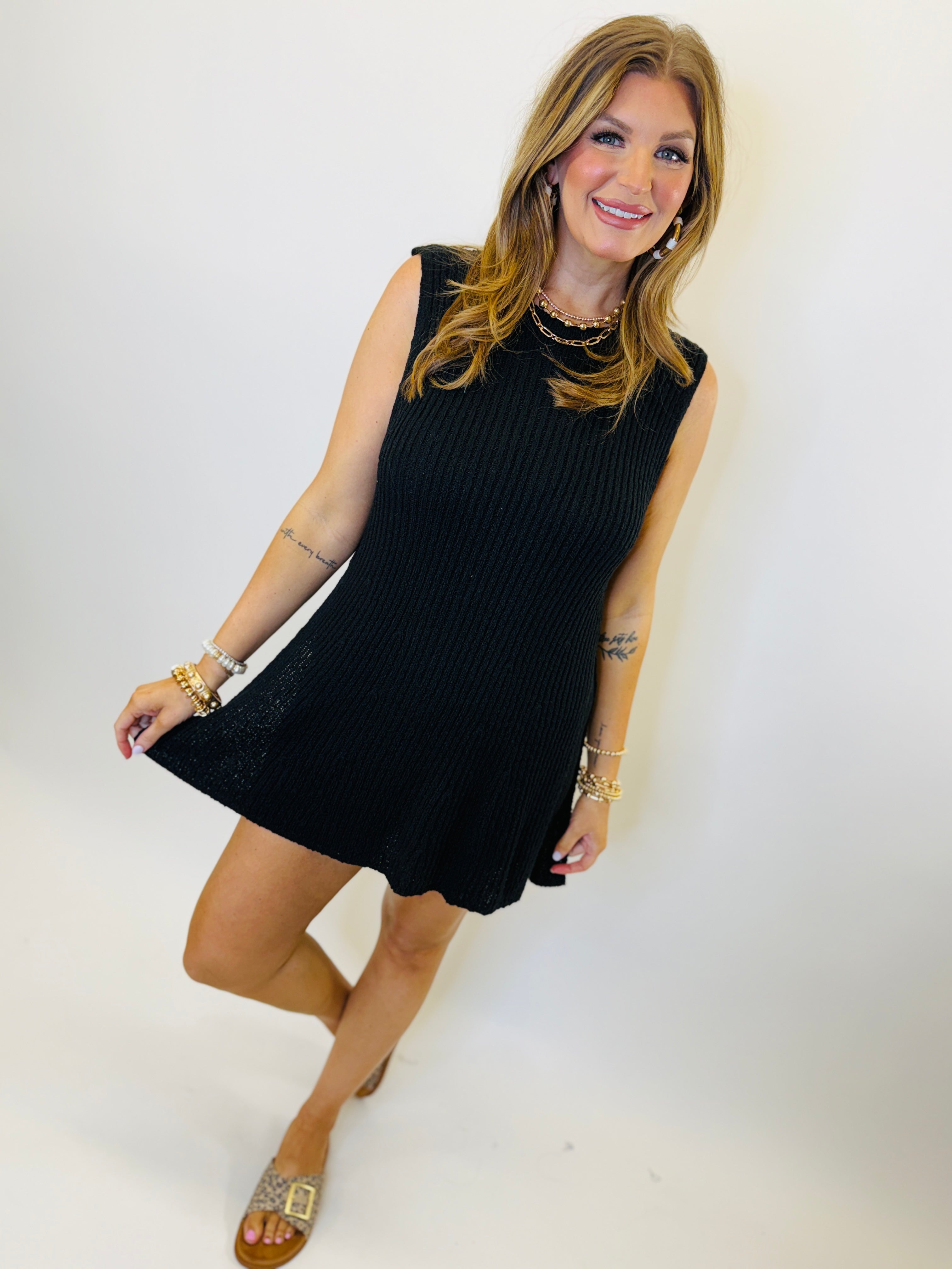 Woman wearing black ribbed sleeveless mini dress with gold jewelry and sandals against white background