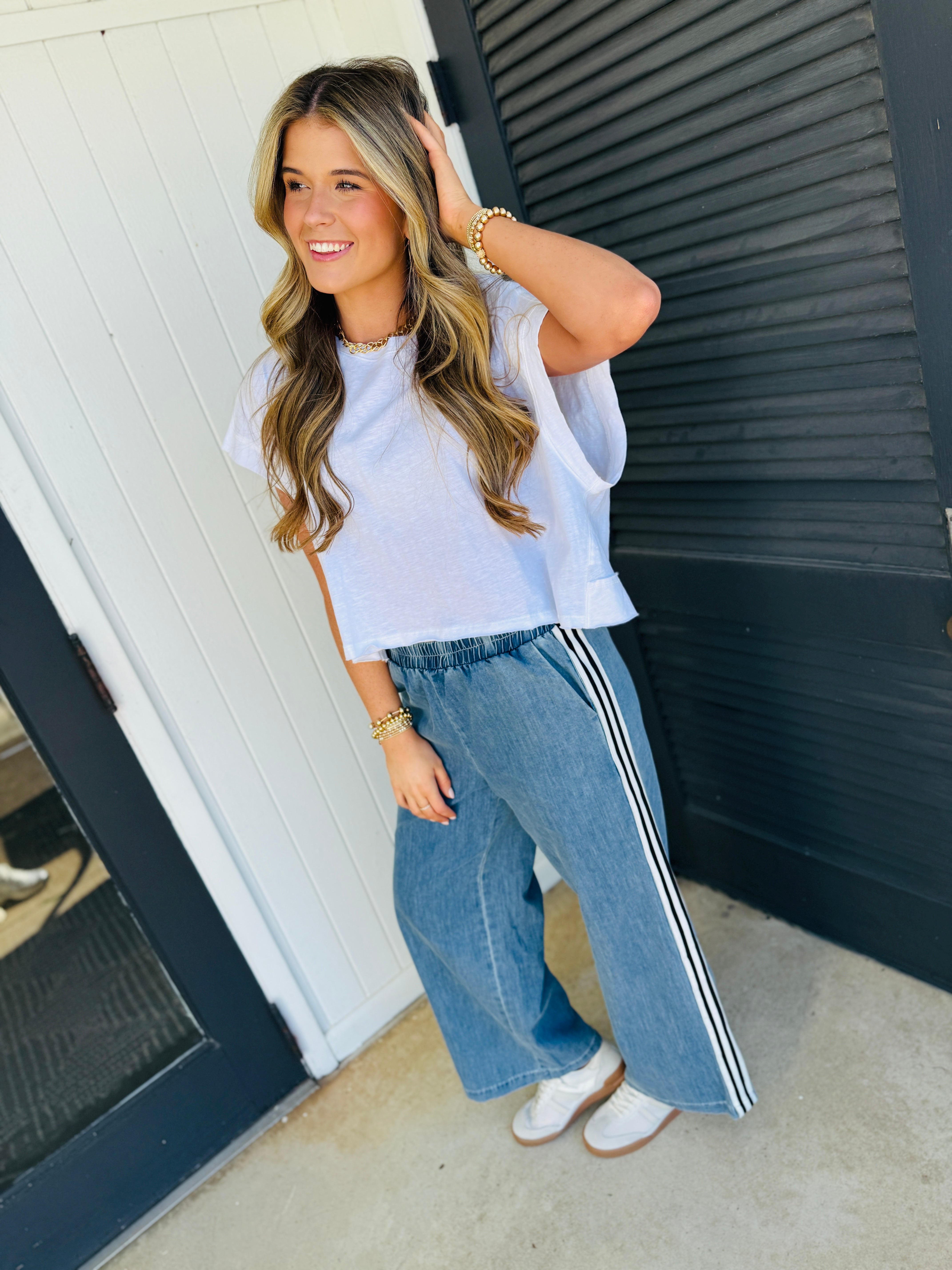Woman wearing relaxed blue denim pants with white side stripes and white top, smiling outdoors.