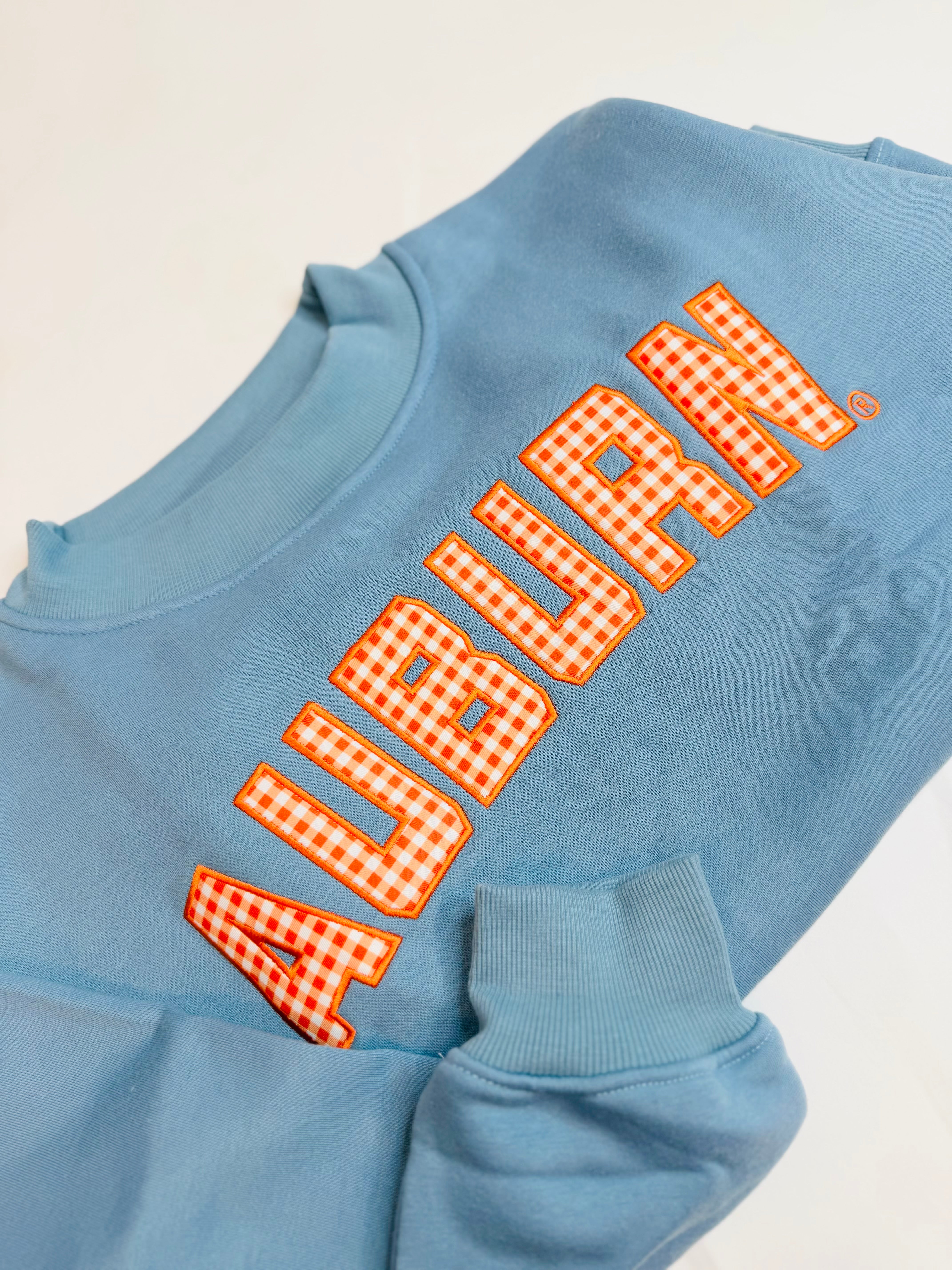 Light blue crewneck sweatshirt featuring orange gingham appliqué "AUBURN" lettering on the chest.