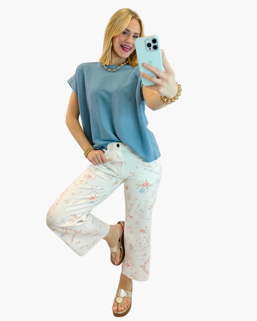 Woman wearing light blue Maya Classic Everyday Top with floral pants, taking mirror selfie while smiling