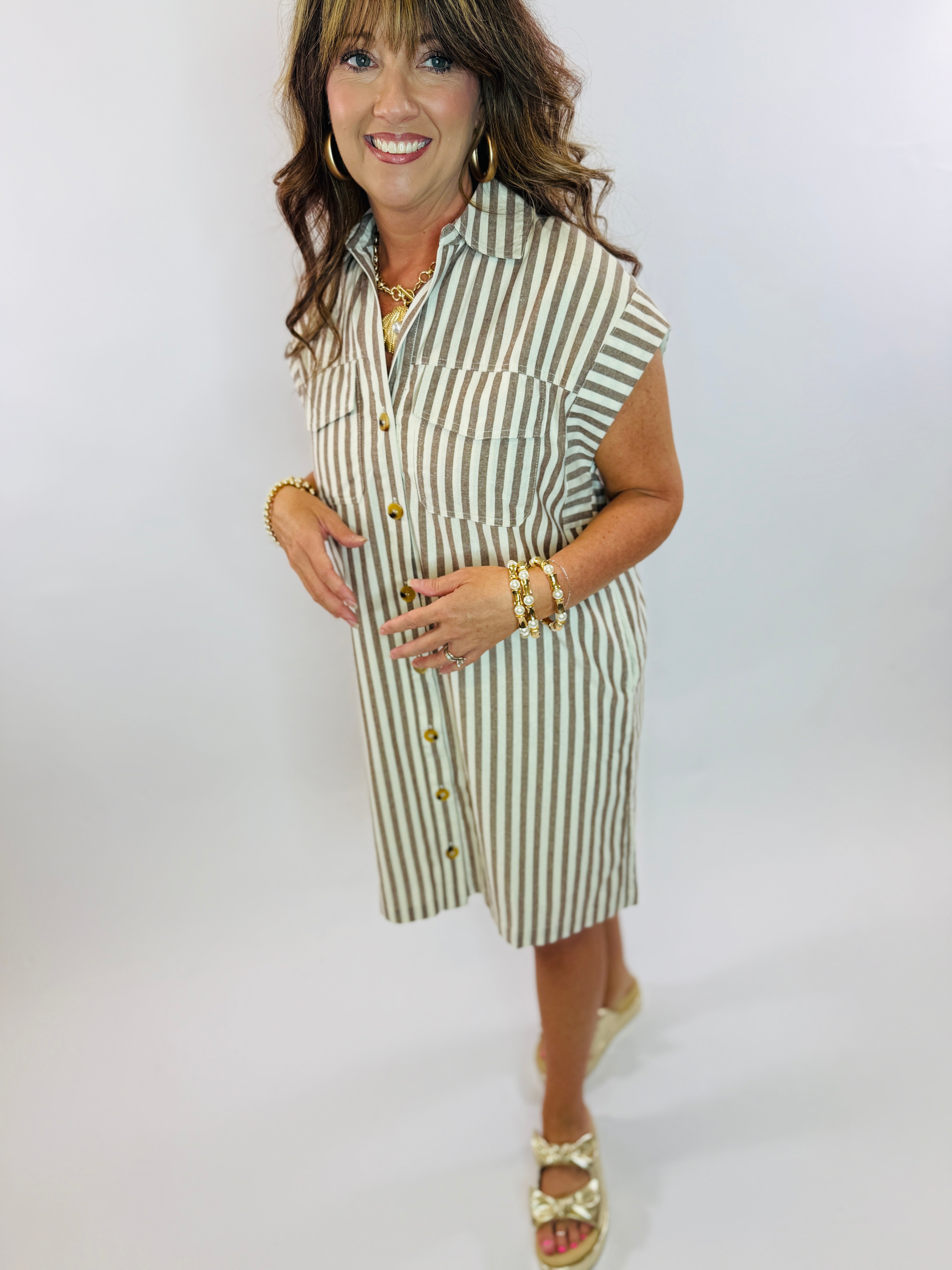 Woman wearing olive and cream striped sleeveless button-front shirt dress with gold accessories and sandals.