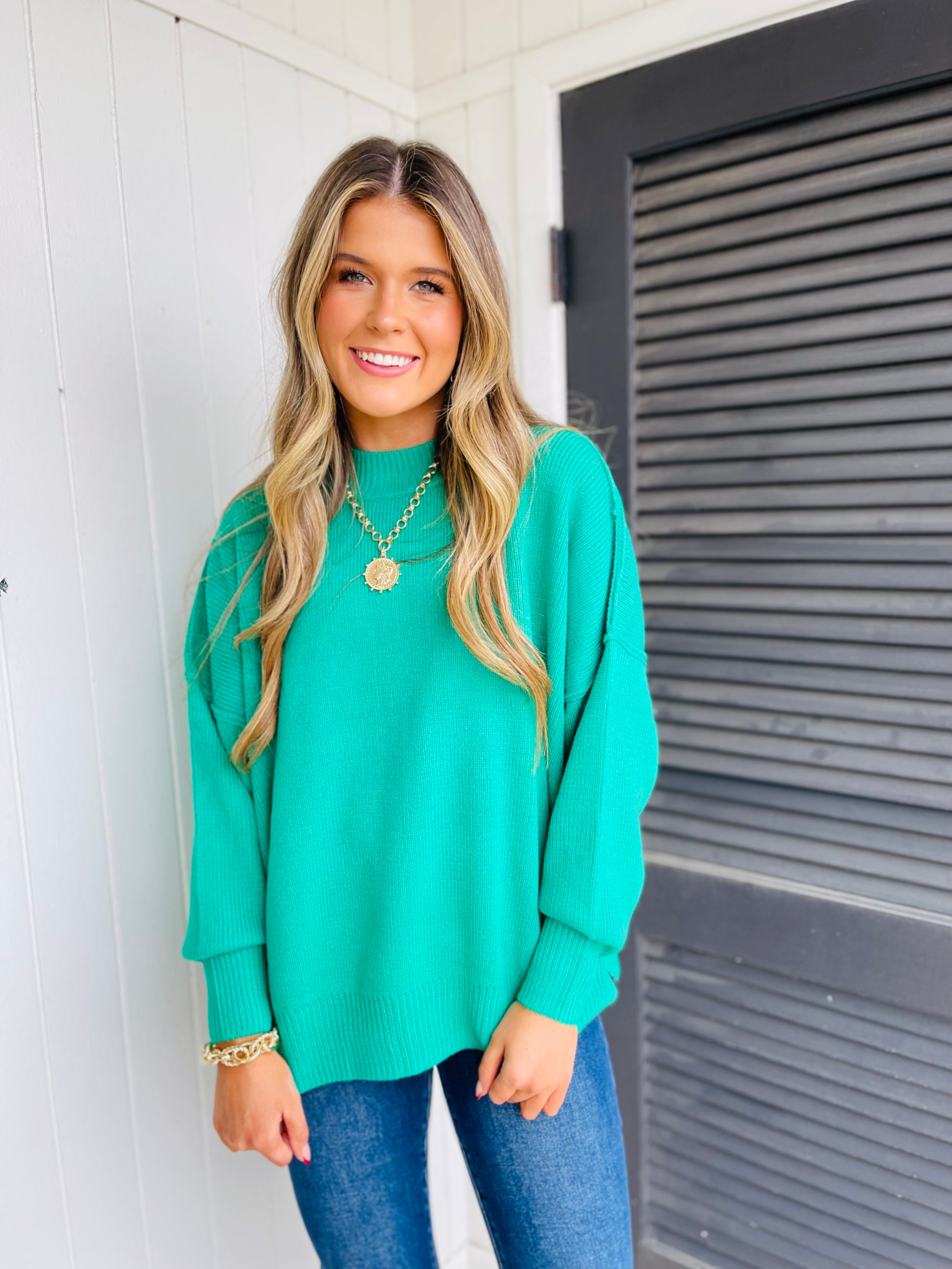Woman wearing bright turquoise oversized knit sweater with gold necklace and blue jeans, smiling outdoors.