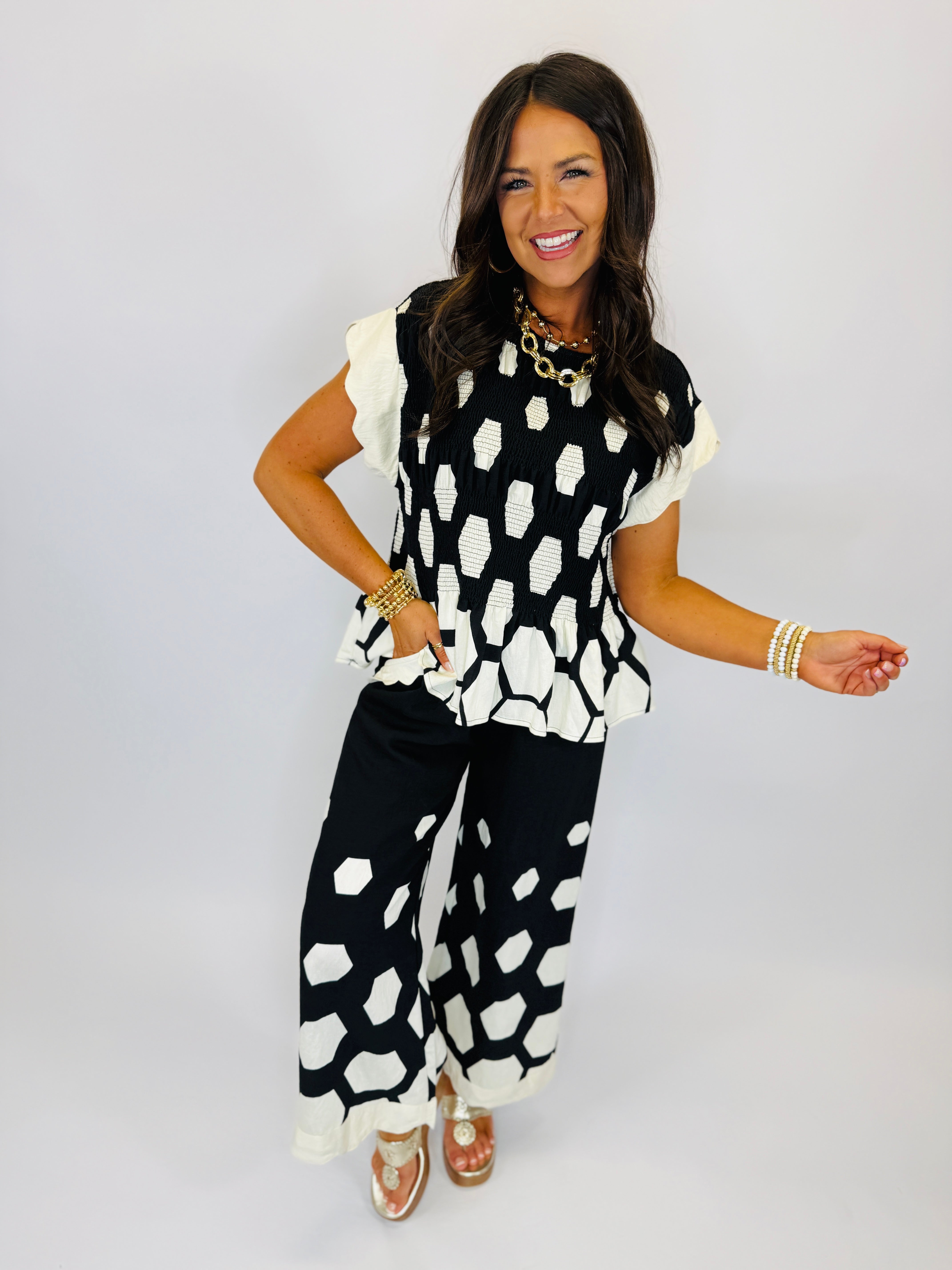Woman wearing black and white polka dot ruffled top with matching wide-leg pants, styled with gold jewelry and sandals.