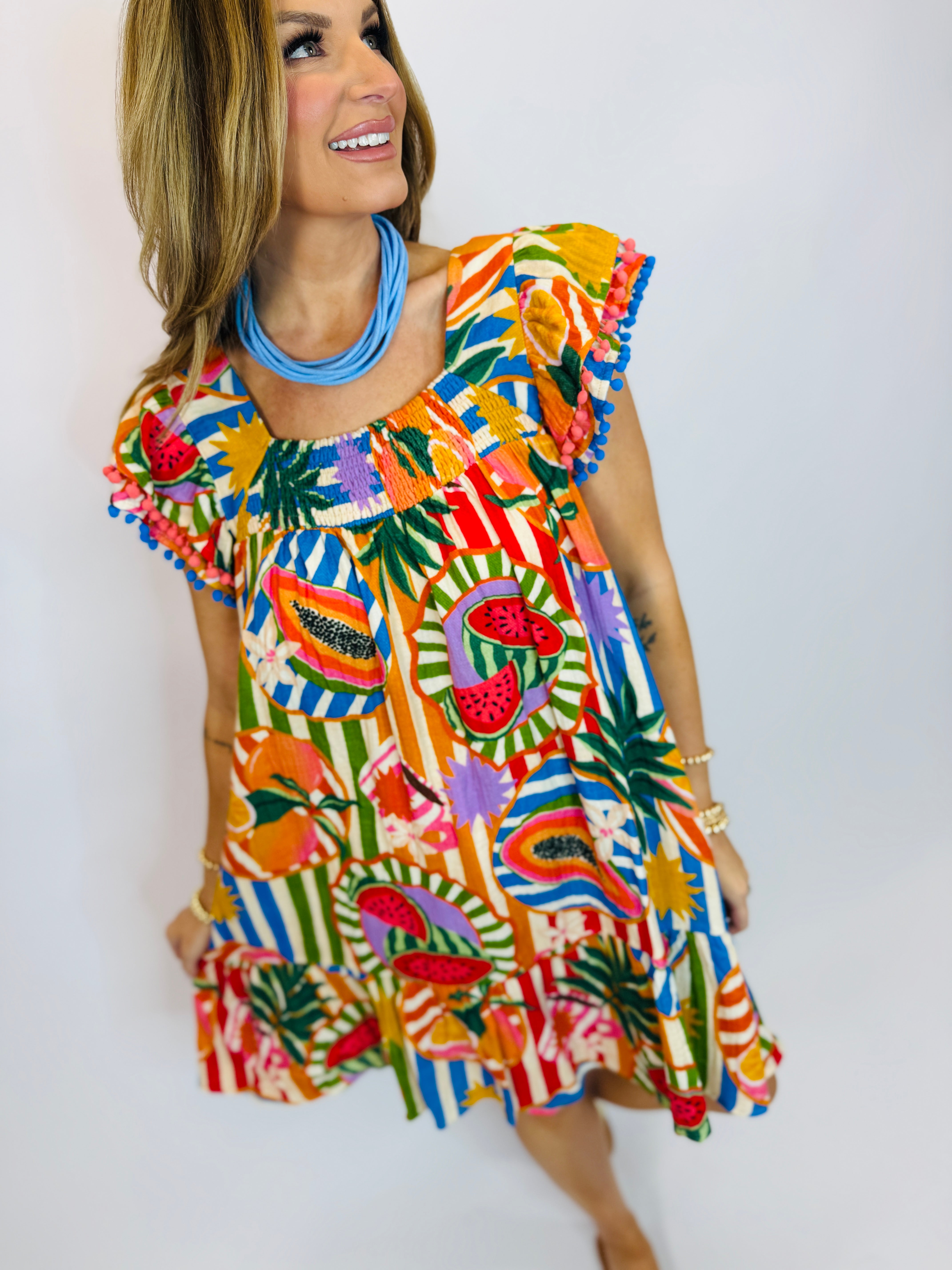 Woman wearing colorful tropical print Luca dress with ruffled shoulders and pom-pom trim, featuring fruit and palm leaf mo...