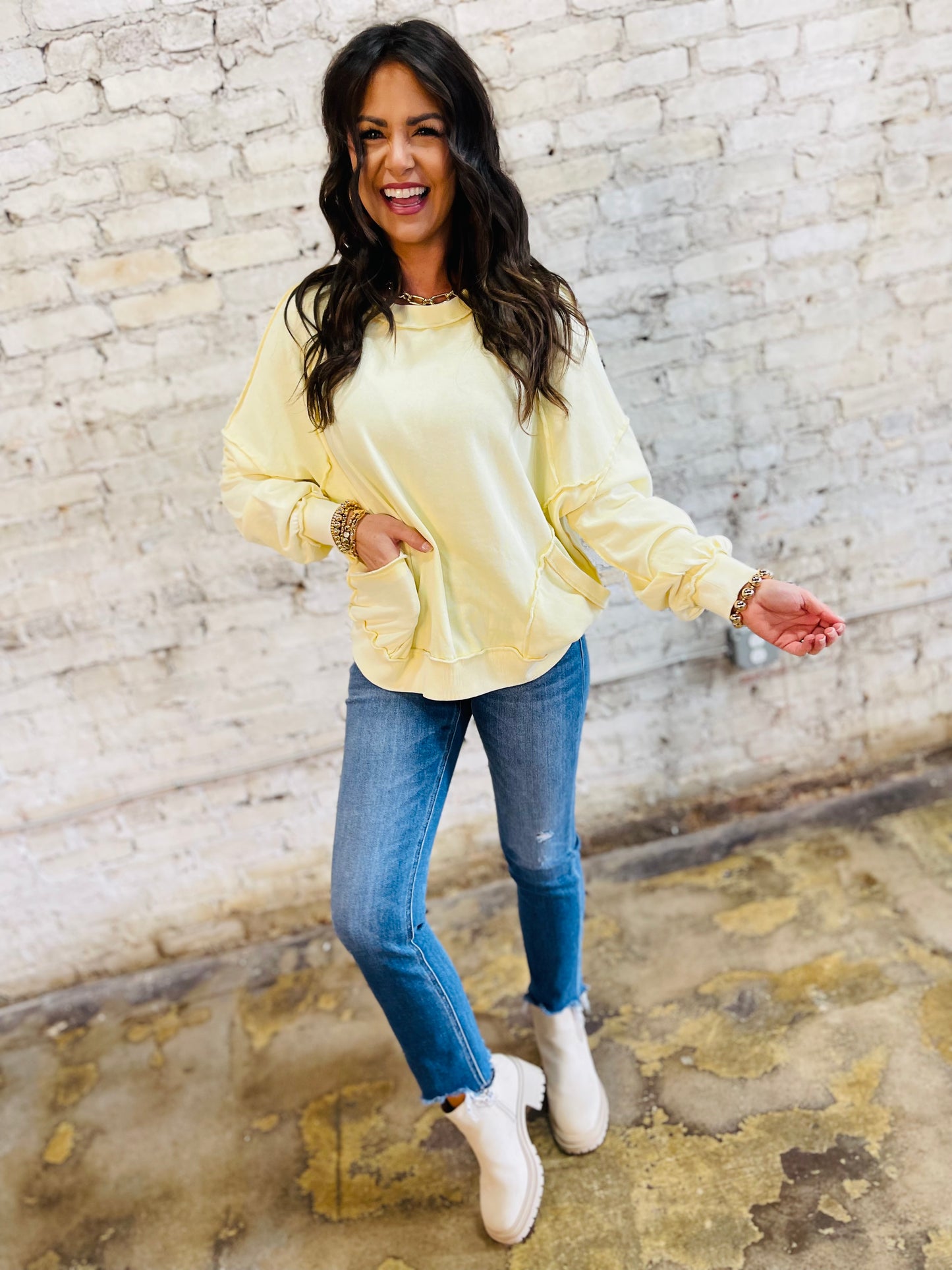 Woman wearing yellow oversized pullover sweatshirt with blue jeans and white sneakers against brick wall