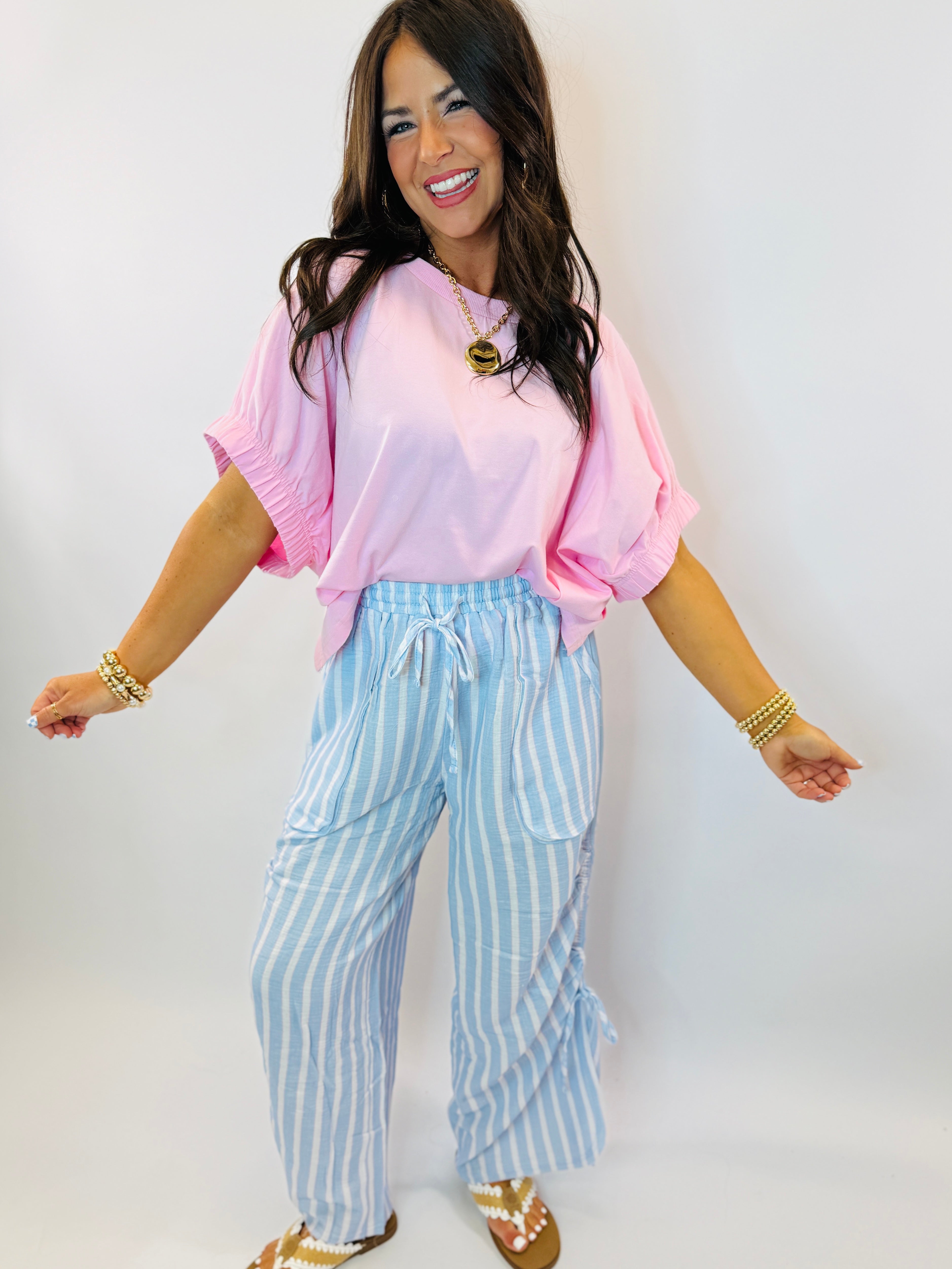 Woman wearing pink oversized Lucy top with ruched sleeves paired with blue striped drawstring pants and gold accessories.