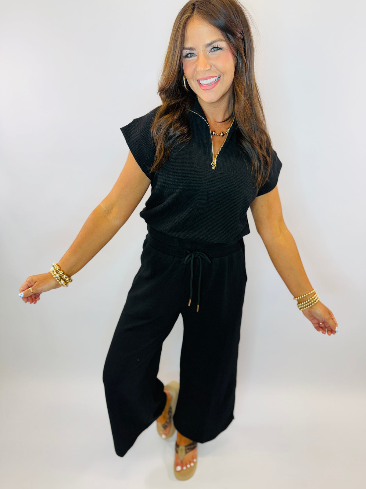Woman wearing black ribbed sleeveless top with zipper and drawstring waist, styled with gold jewelry and wide-leg pants.