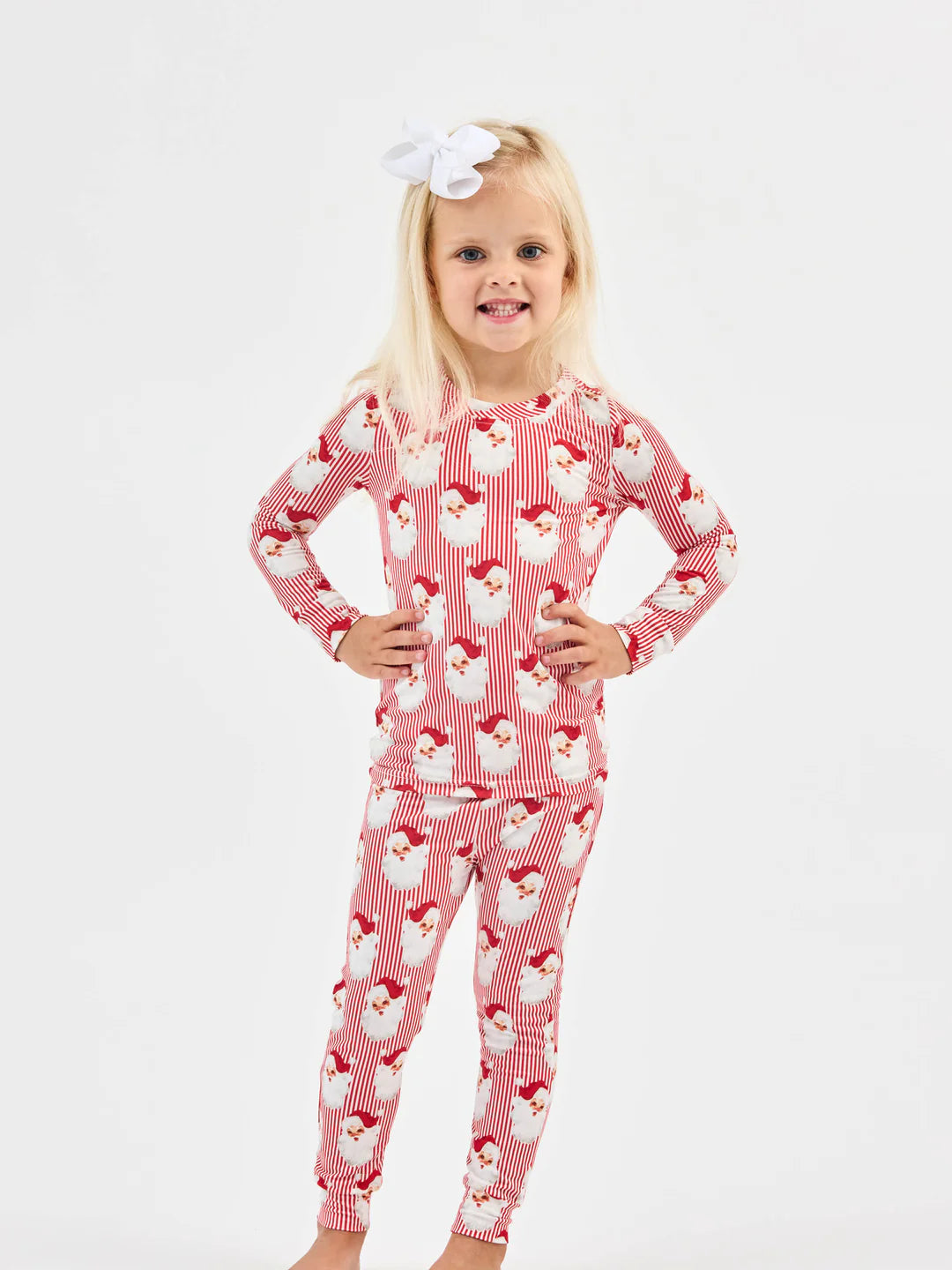Blonde toddler girl wearing red and white striped Santa-patterned Christmas pajama set with white bow headband