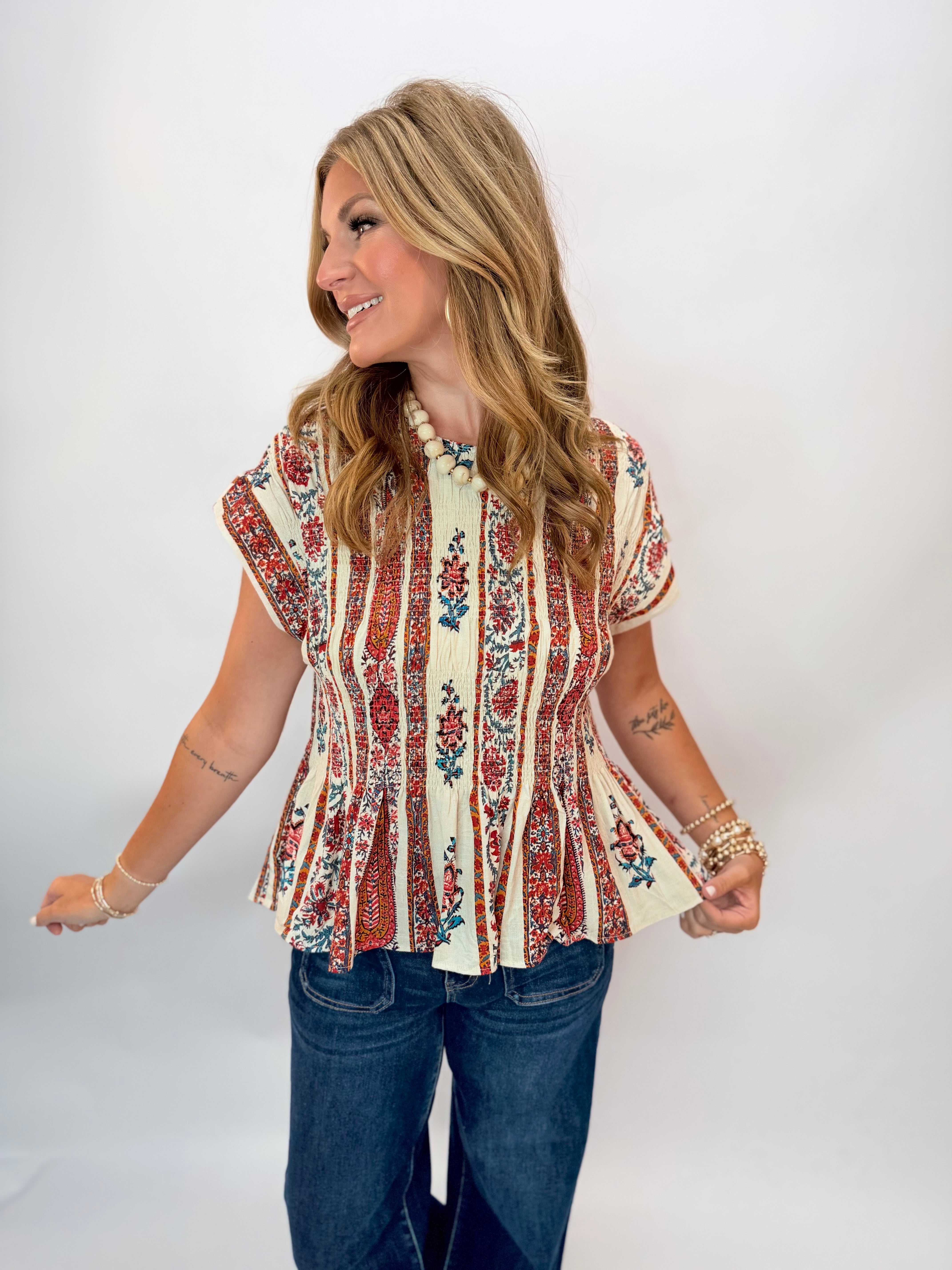 Woman wearing the Renee Top, a cream floral-print smocked peplum blouse with short sleeves, paired with dark jeans.